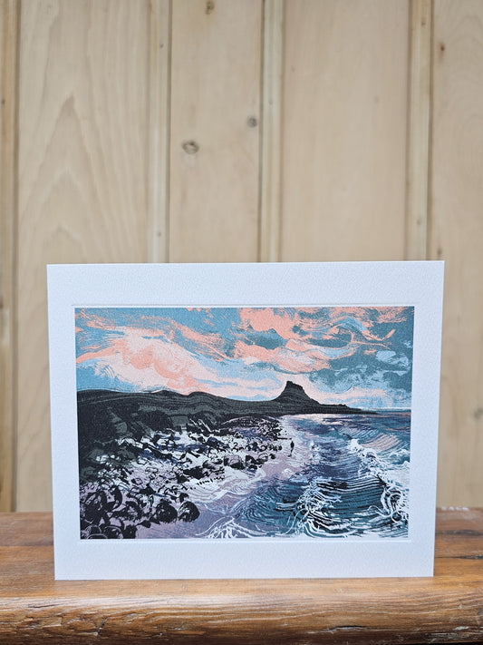 Greeting card reproducing Andy Lovell's linocut print Early Morning Light, showing a lone figure on a rocky shore with rolling waves and a distinctive rocky headland beneath a dramatic pink and blue dawn sky
