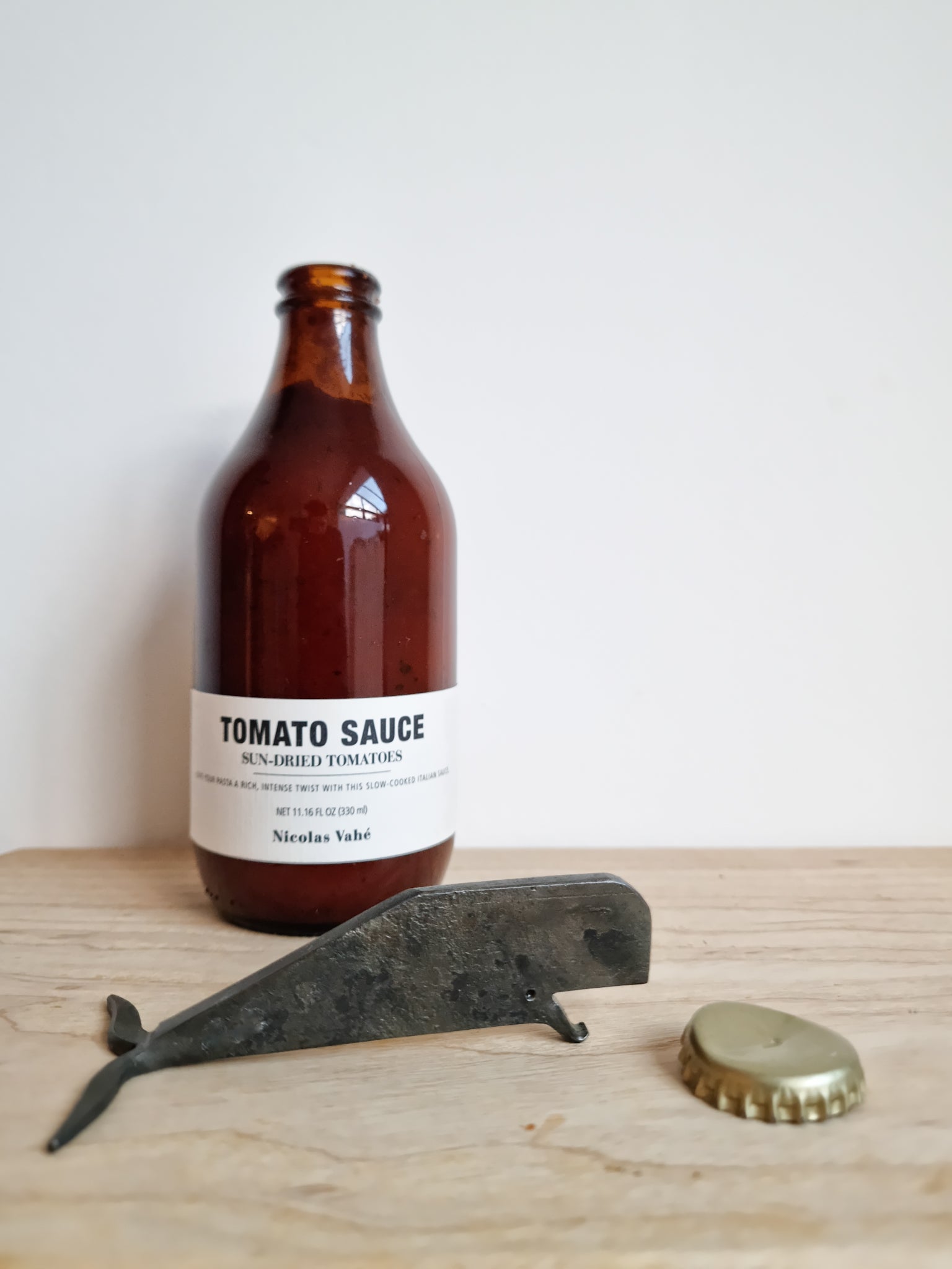 A hand forged iron bottle opener shaped like a whale, placed on a wooden surface with a brown bottle labeled 'Tomato Sauce' in the background and a copper bottle cap beside the opener.