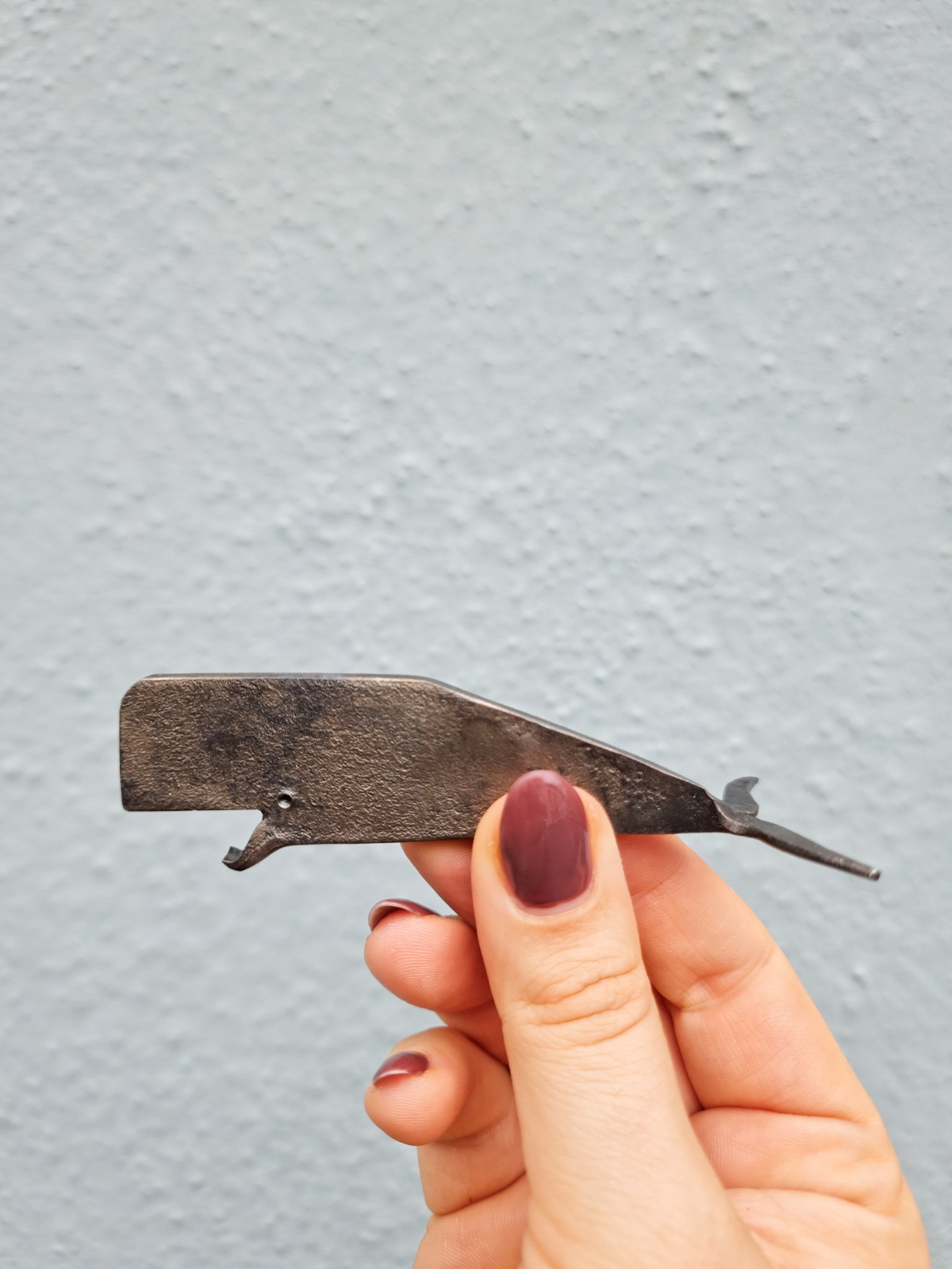 A hand forged iron bottle opener shaped like a whale, held up against a blue background. 