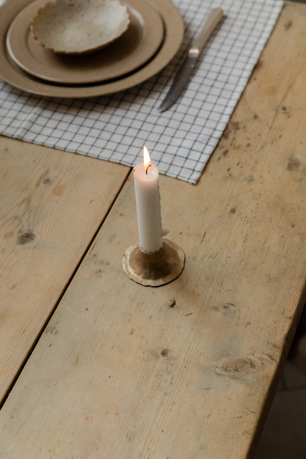 Lit white candle in a hand-forged antiqued brass holder with tulip base beside a place setting on a grid linen placemat