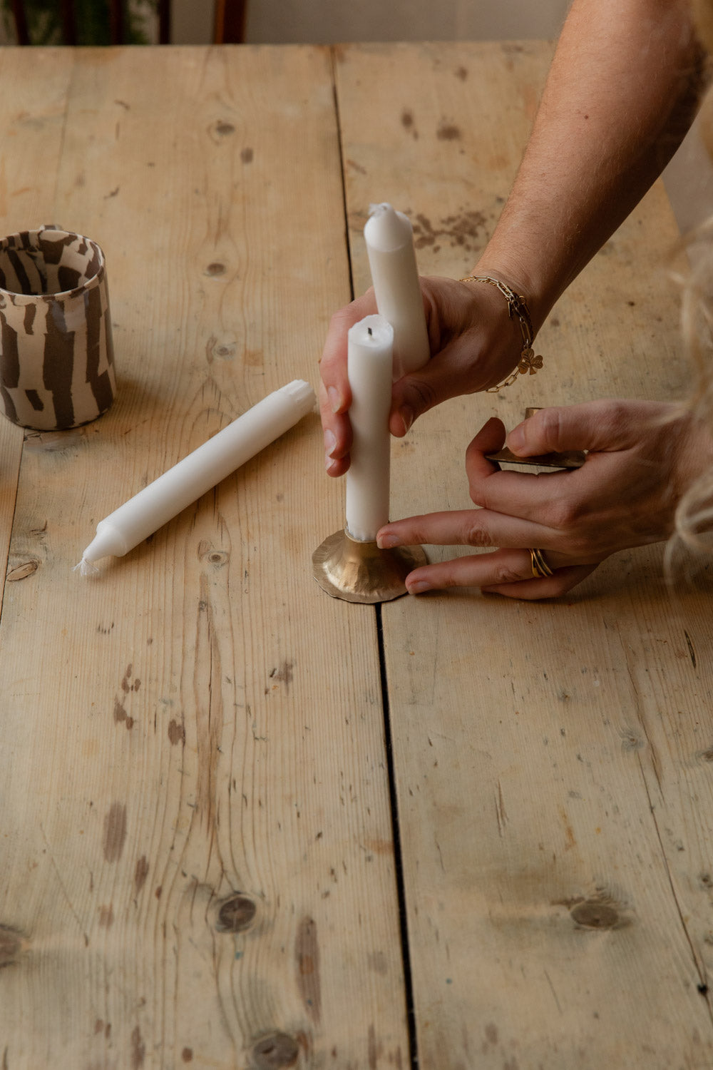 Hands fitting a white dinner candle into a hand-forged antiqued brass candle holder on a pine table