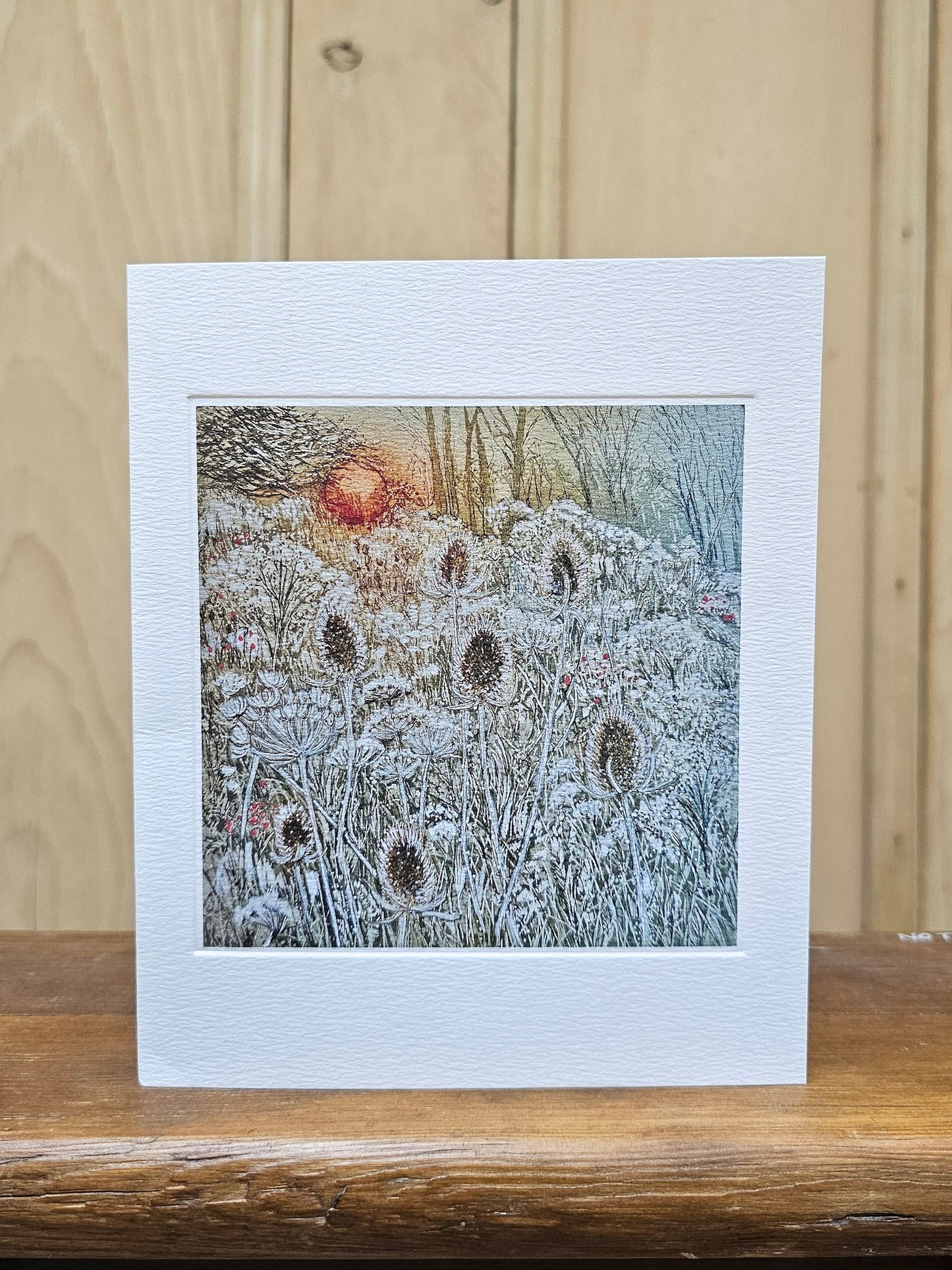 A rectangular greeting card with a wide white border, featuring a winter landscape illustration of frosty wildflowers and bare trees under a glowing orange sun. The card is displayed standing upright on a wooden surface against a light wood paneled background.