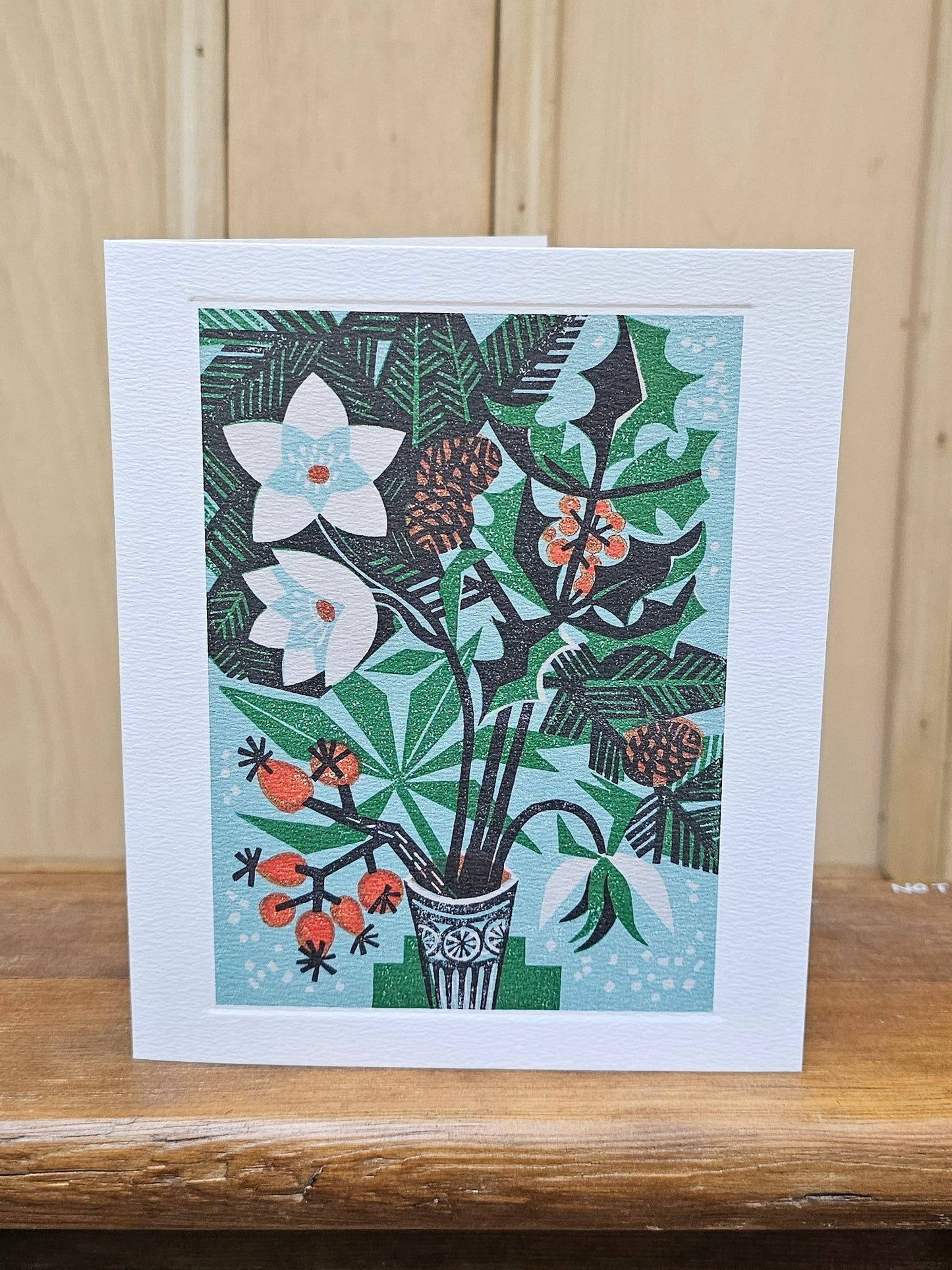 A rectangular white greeting card with a colorful botanical illustration of flowers, leaves, and berries in a vase, standing on a wooden surface against a light wood paneled background.