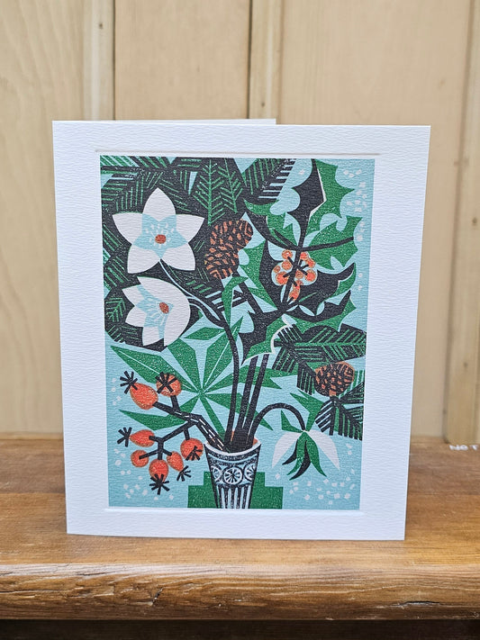 A rectangular white greeting card with a colorful botanical illustration of flowers, leaves, and berries in a vase, standing on a wooden surface against a light wood paneled background.
