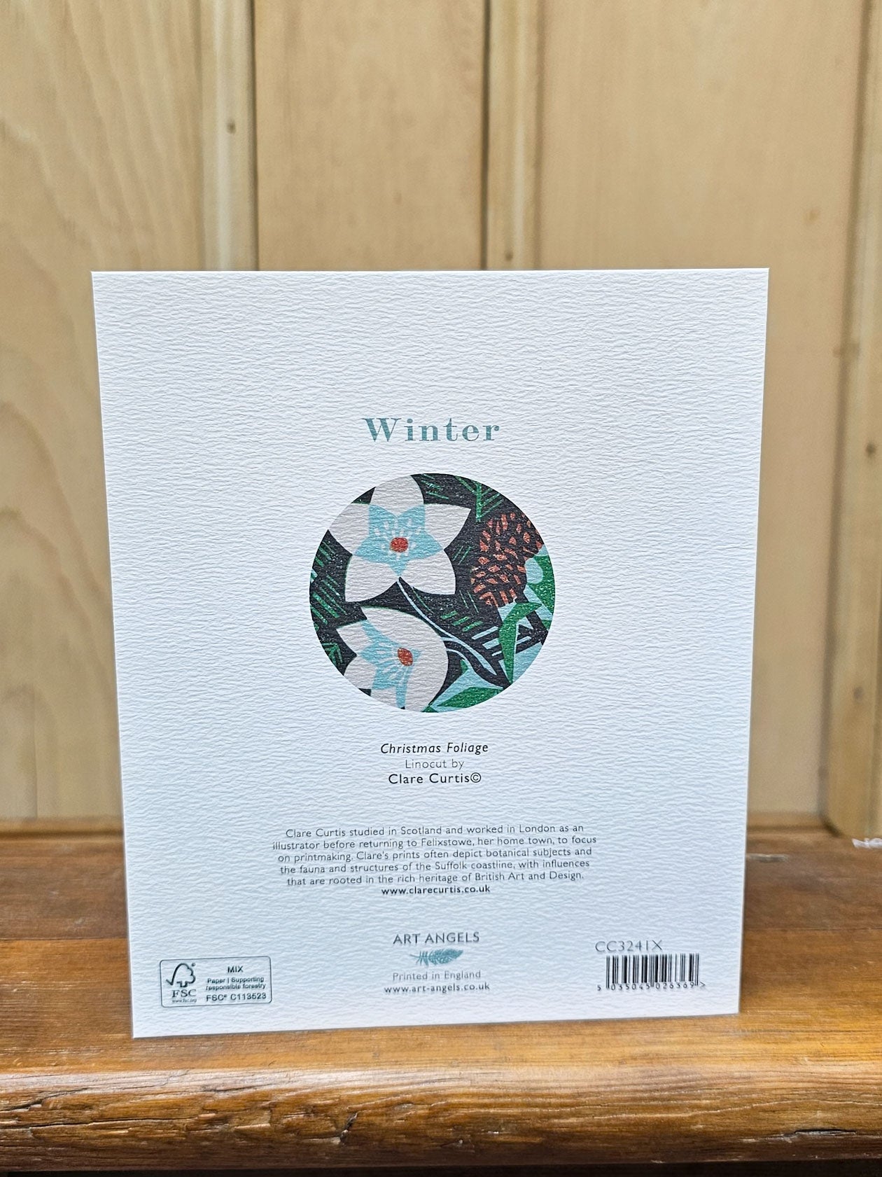 The back of a textured white card of Winter Christmas Felage, featuring a circular floral illustration and product details, set against a wooden background.