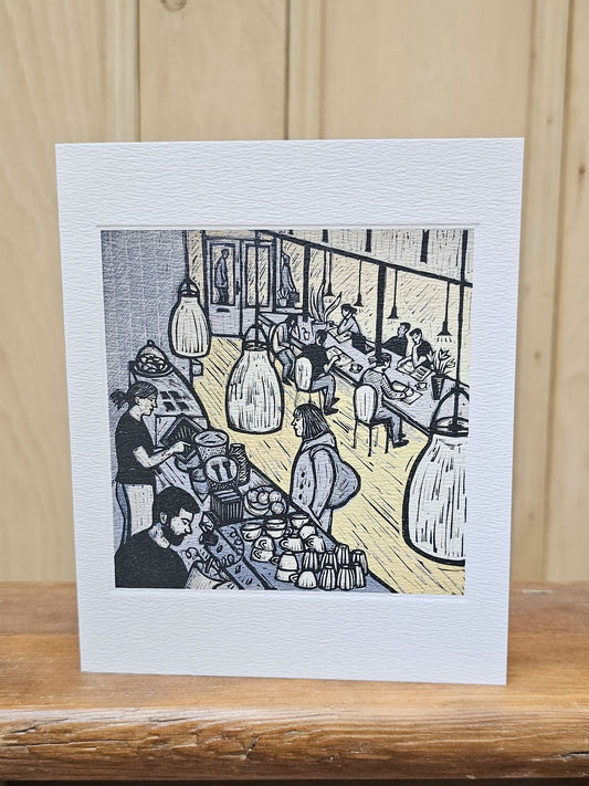 A rectangular art print card with a wide white border featuring a black, gray, and yellow linocut-style illustration of a bustling cafe interior, displayed on a wooden surface against a light wood background.