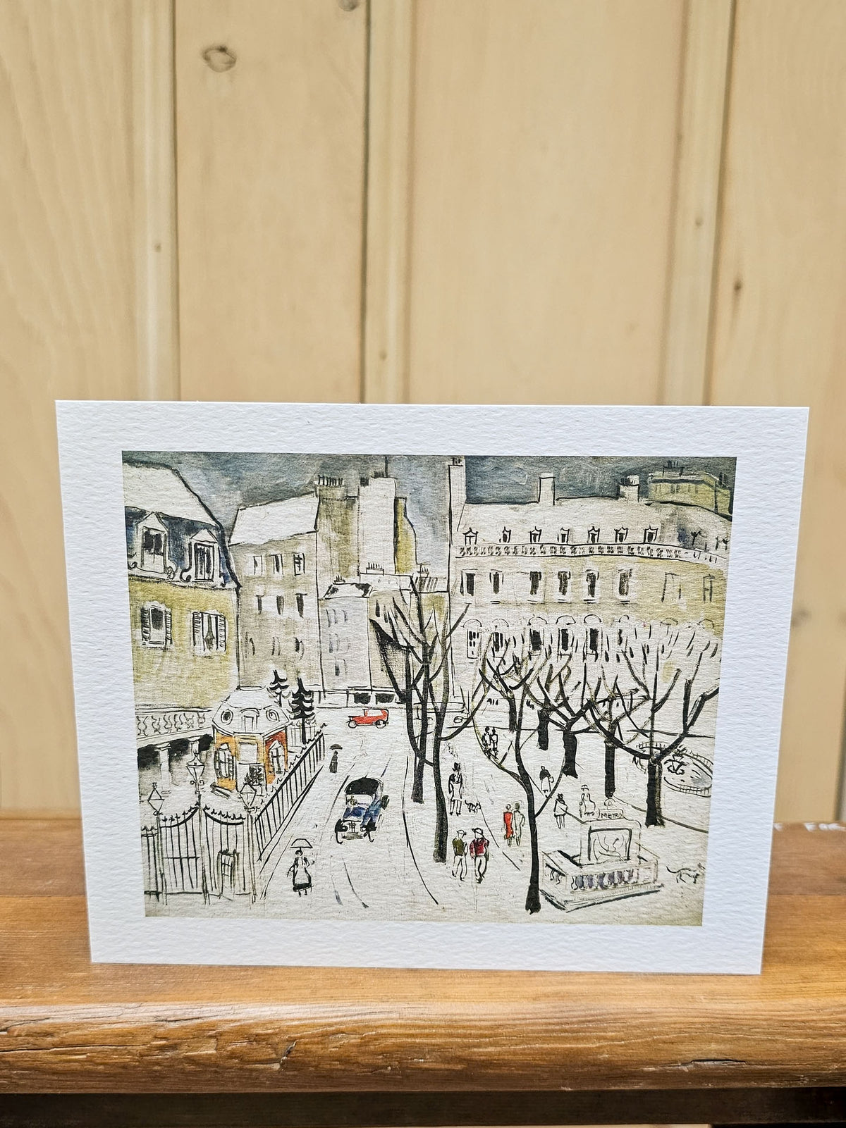 A rectangular art print card featuring a hand-drawn winter Paris street scene with buildings, leafless trees, and small figures, displayed on a wooden surface against a light wood plank background.