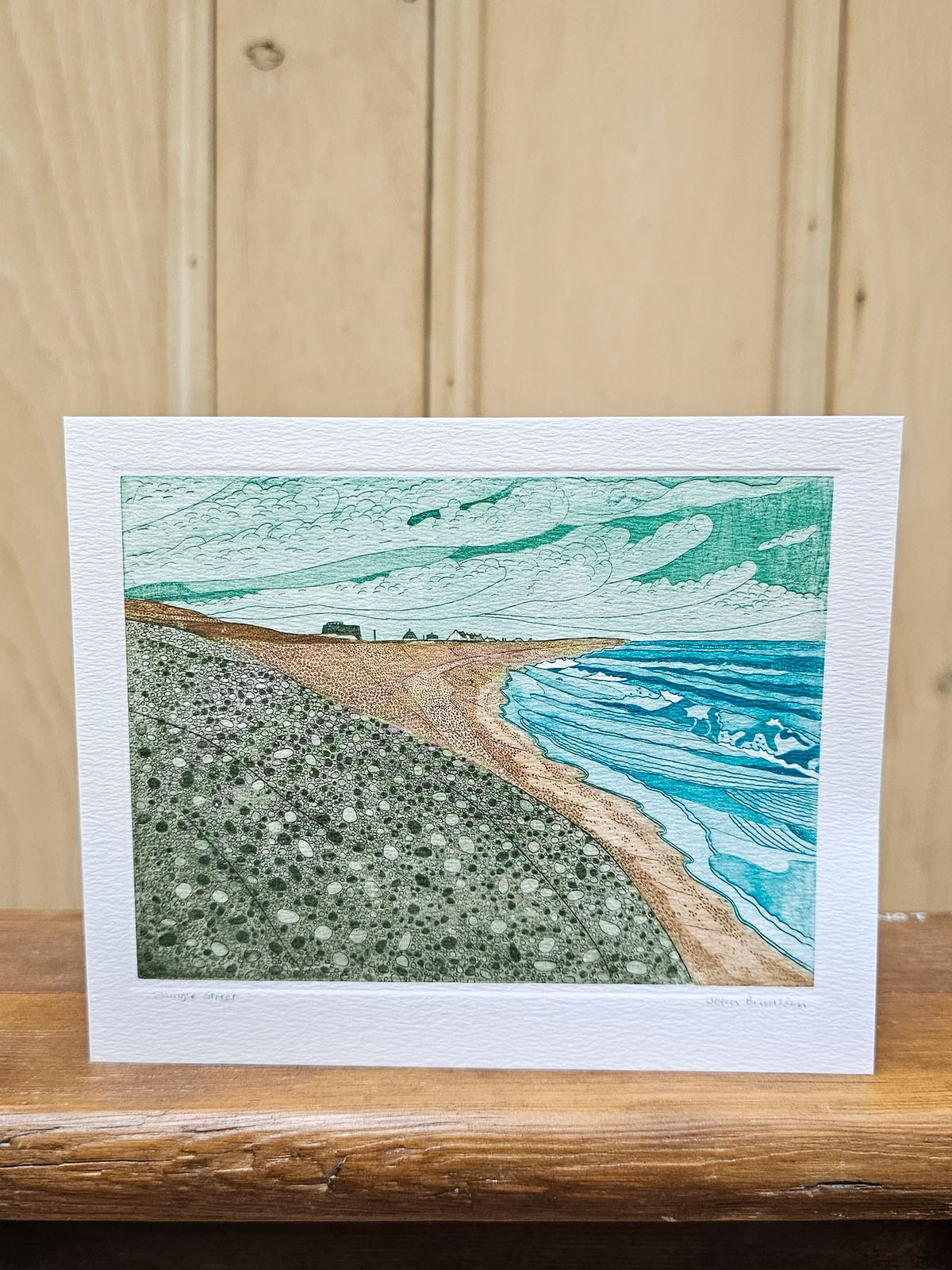 A rectangular art print card depicting a coastal landscape with turquoise ocean waves, a sandy beach, and a textured green shoreline, displayed on a wooden surface against a light wood paneled background.