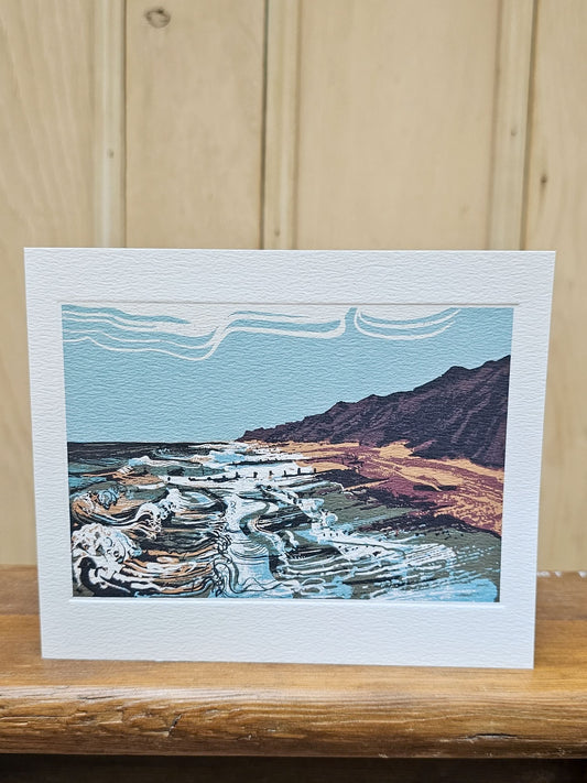 A square rectangular art print greetings card with a wide white border, featuring a stylized landscape illustration of waves, a beach, and mountains in blue, brown, beige, and white, displayed on a wooden surface against a light wood plank background.