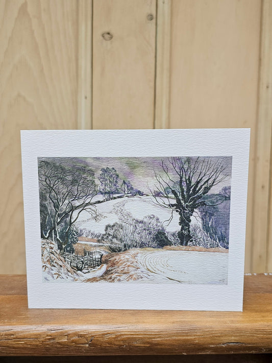 A rectangular art print greetings card with a wide white border showing a winter landscape painting, displayed on a wooden surface against a light wood paneled background.