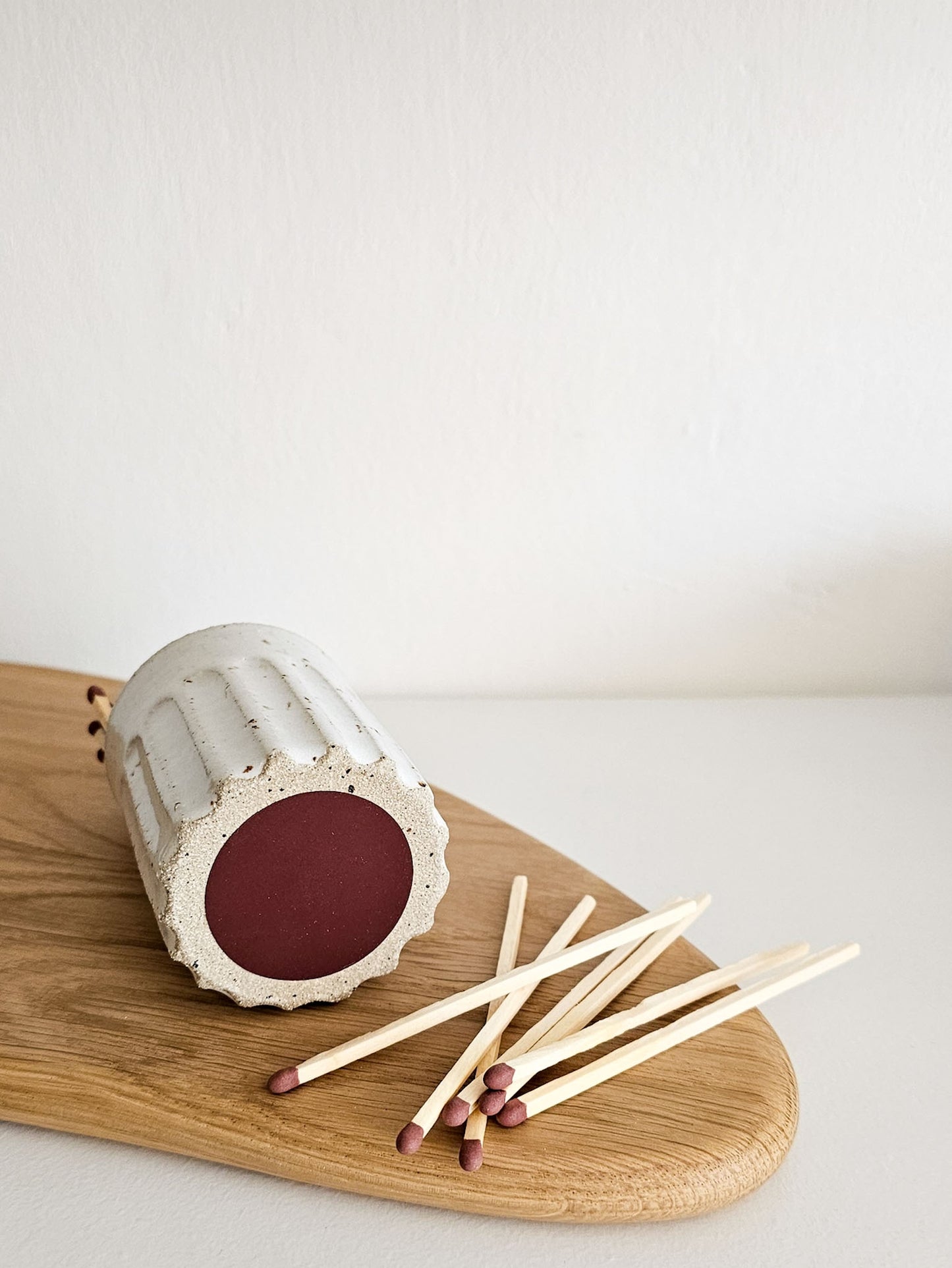 A ceramic matchbox with a ribbed cylindrical body and a dark red circular end, shown lying on a wooden board with several wooden matches beside it against a plain light background.