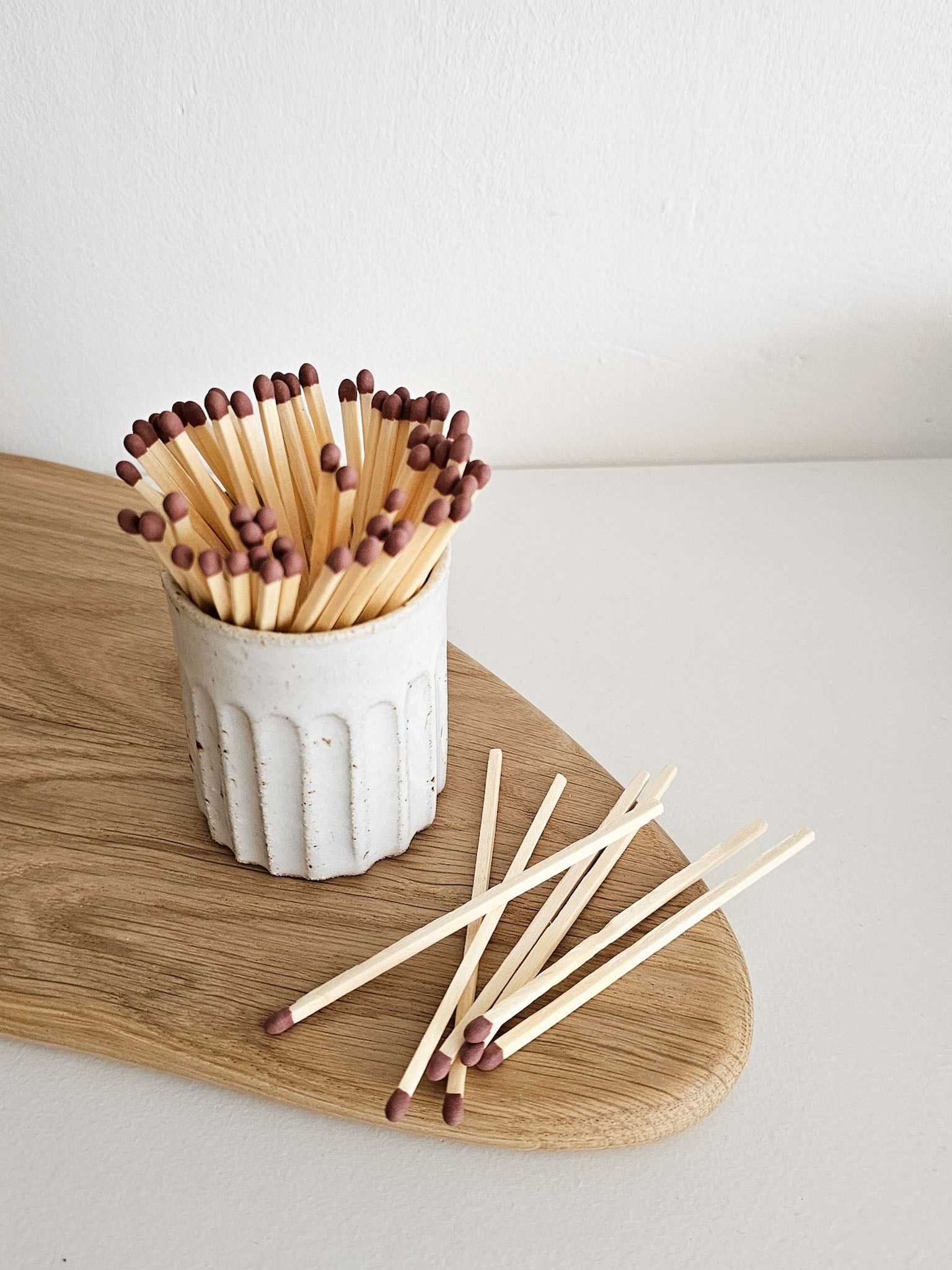 A small white ribbed ceramic container filled with wooden matchsticks with dark brown tips, placed on a light wood board against a plain off-white background.