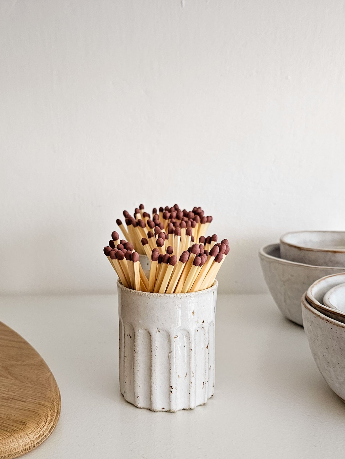 A white speckled ceramic match holder filled with wooden matches with dark tips, set on a light tabletop with bowls in the background.