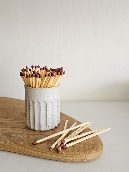 A ceramic match holder filled with wooden matchsticks with dark tips, set on a wooden board against a plain light background.