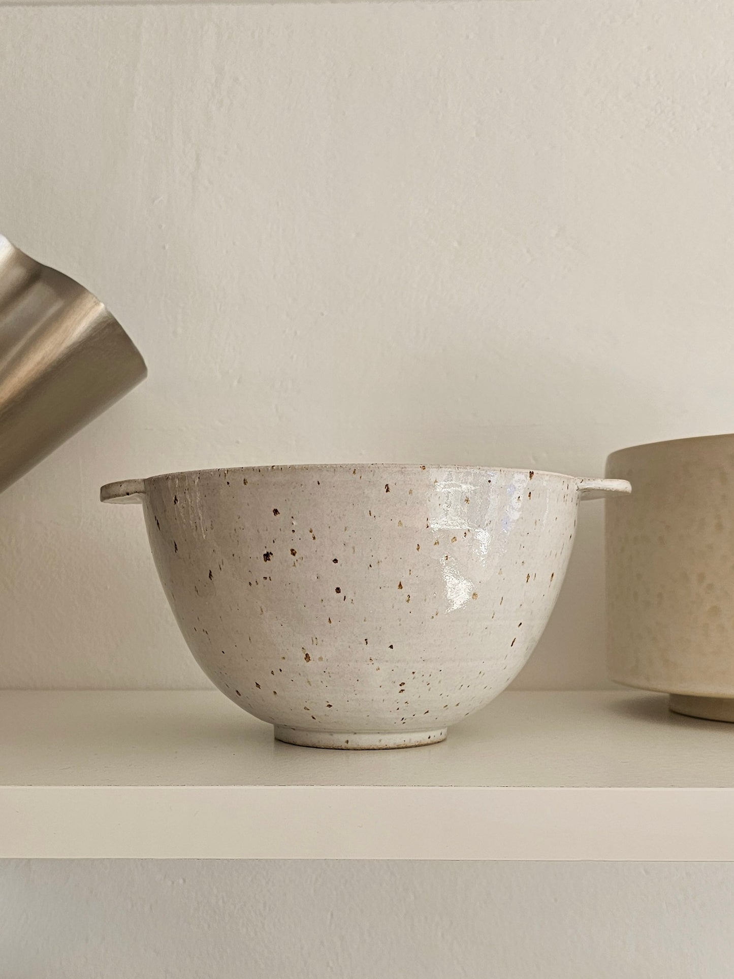 A speckled ceramic bowl with small side handles, shown on a shelf against a neutral wall. Part of a metallic pitcher is visible on the left and another vessel on the right.