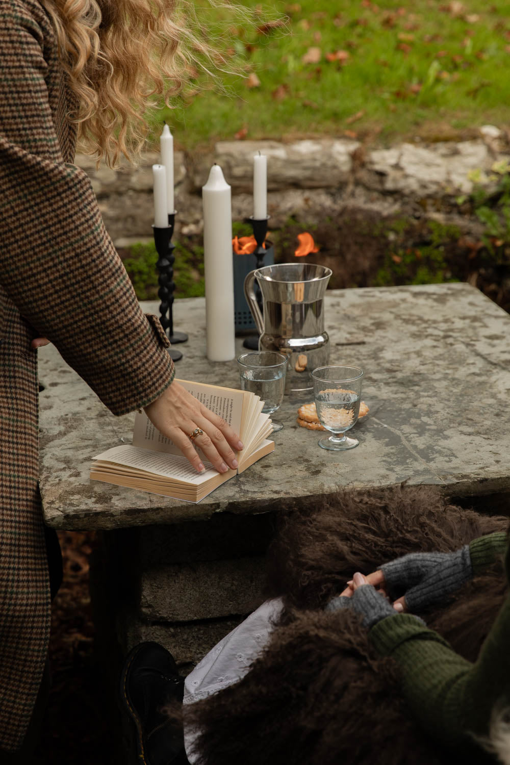 Woman turning a book's pages at a stone garden table set with tall black iron candle holders and white candles