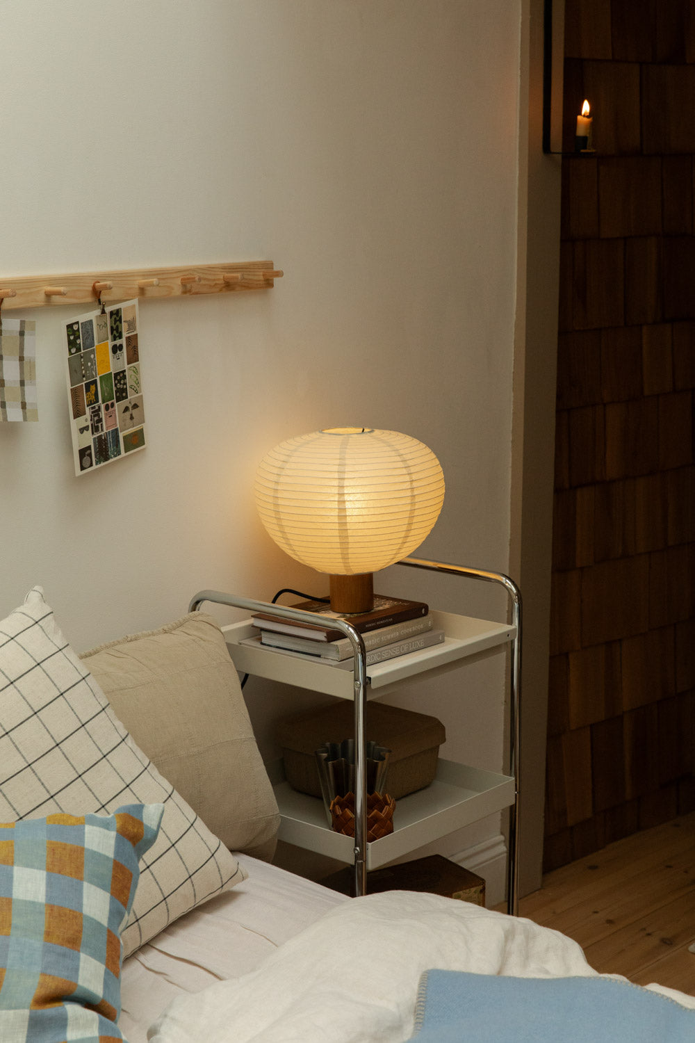 Blomus Kami table lamp glowing on chrome trolley beside bed layered with Klippan Pop lambswool blanket in blue and brown and grid check cushion.