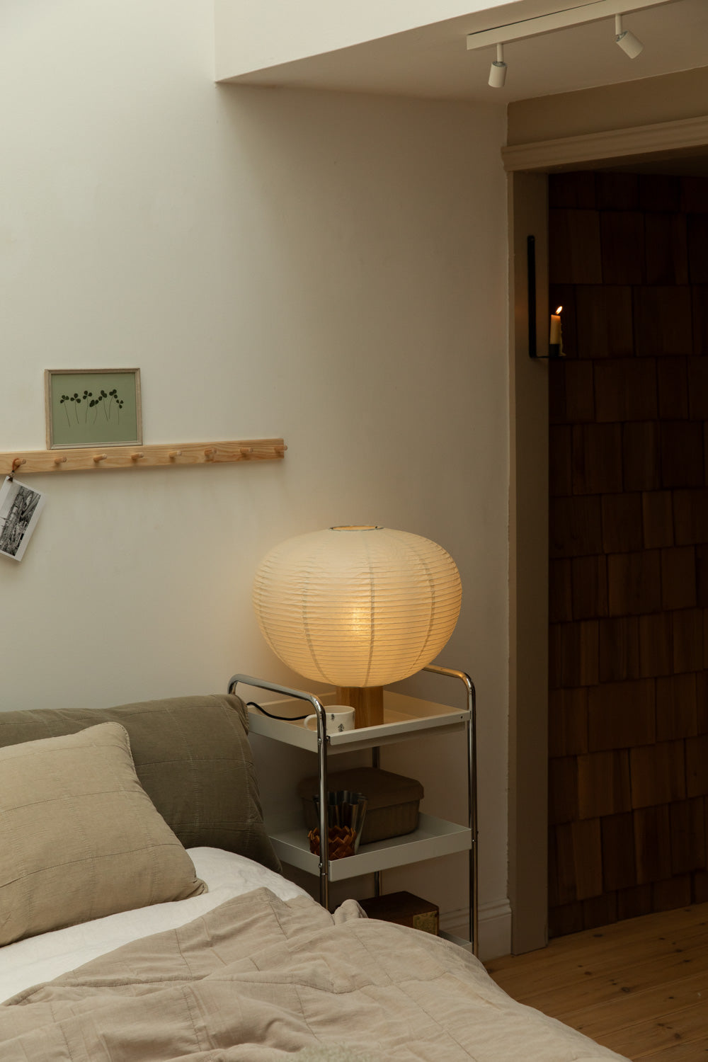 Blomus Kami table lamp on chrome trolley beside olive and vintage linen cushion bed, wall candle sconce glowing in timber-clad doorway, pine peg rail above.