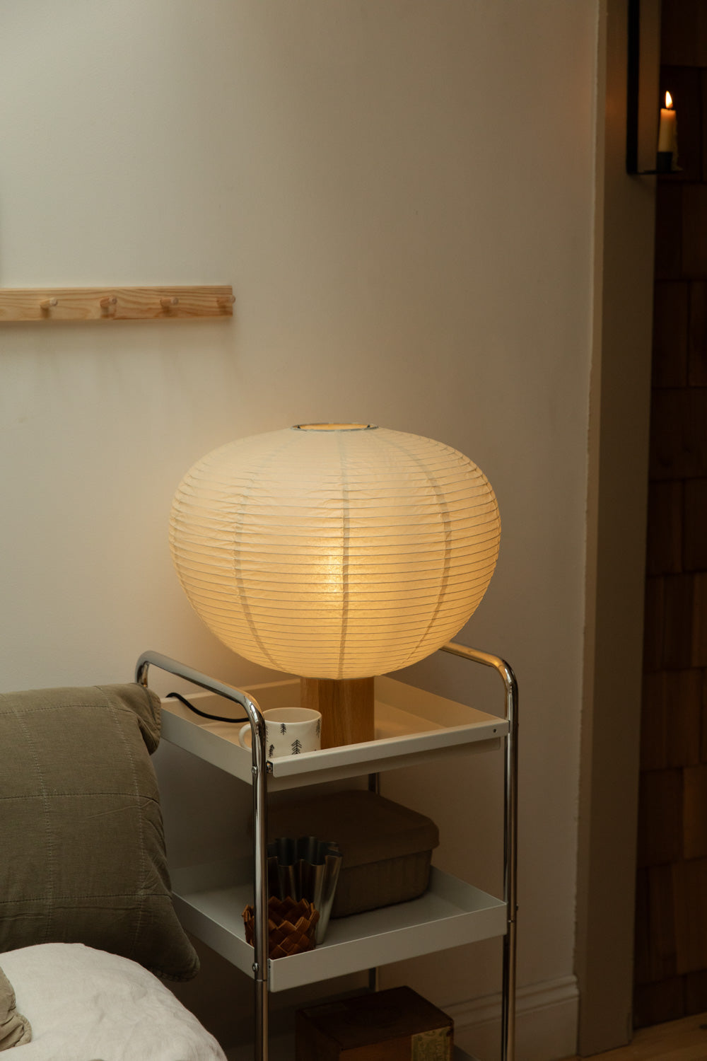 Blomus Kami table lamp on chrome trolley beside olive and vintage linen cushion bed, wall candle sconce glowing in timber-clad doorway, pine peg rail above.