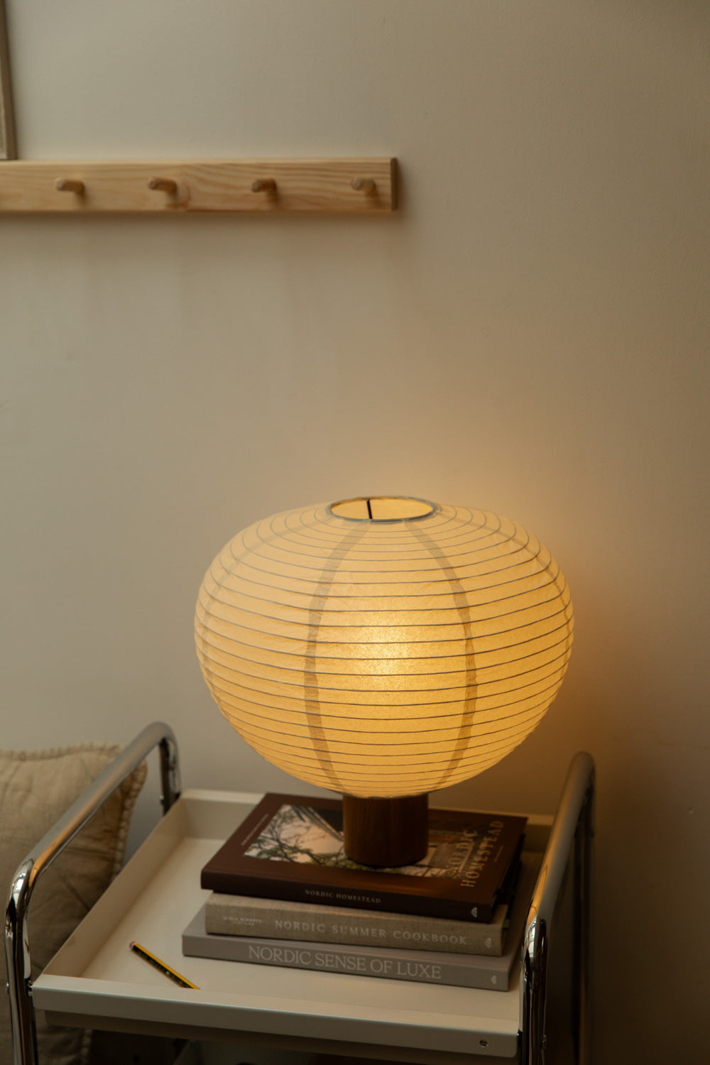Blomus Kami table lamp glowing warmly on oak base, stacked on Nordic books on a chrome trolley nightstand with pine peg rail above.