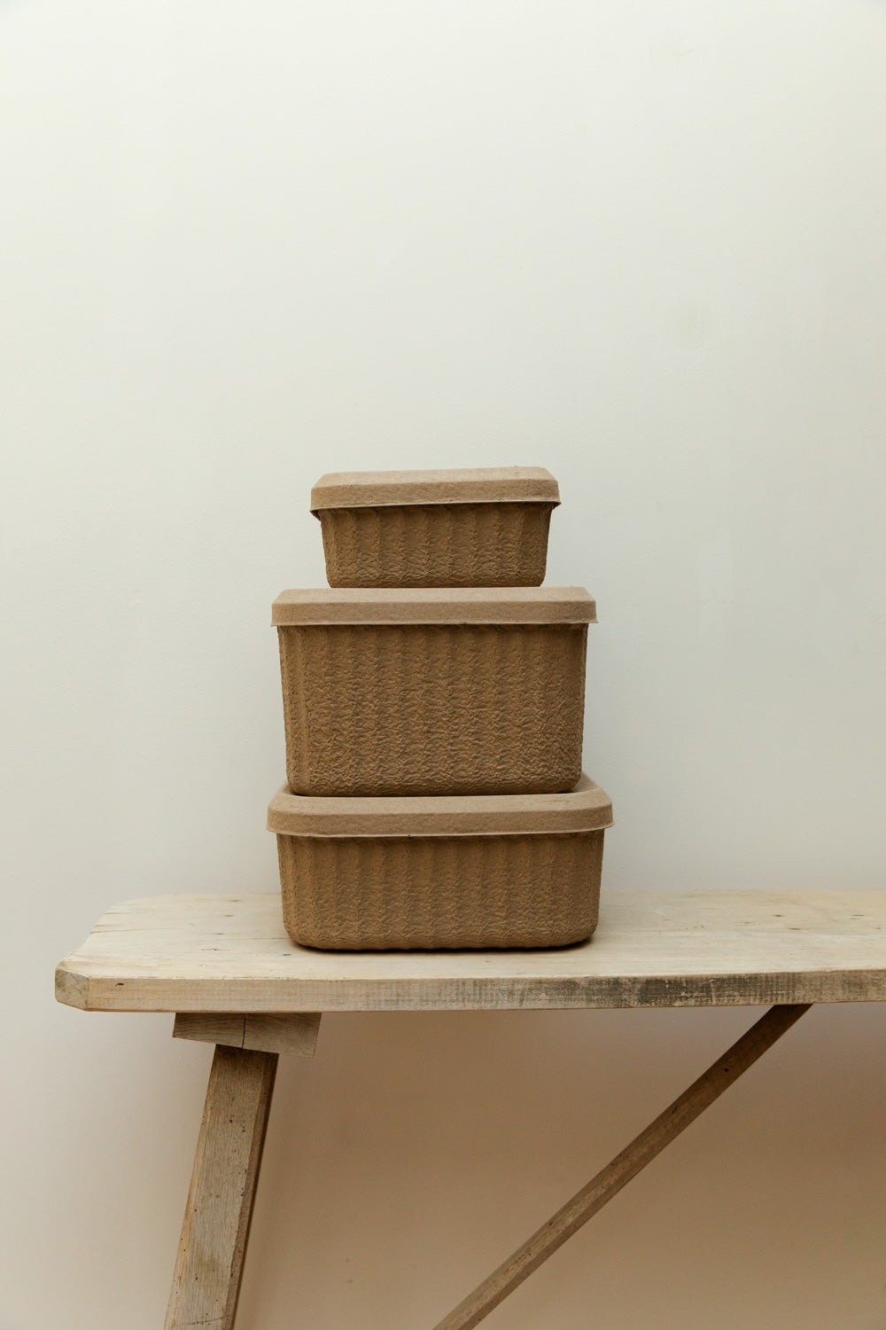 Three Blomus Redo paper pulp storage boxes in small, medium and large stacked in a stepped arrangement on a rustic pale wood table, side view.
