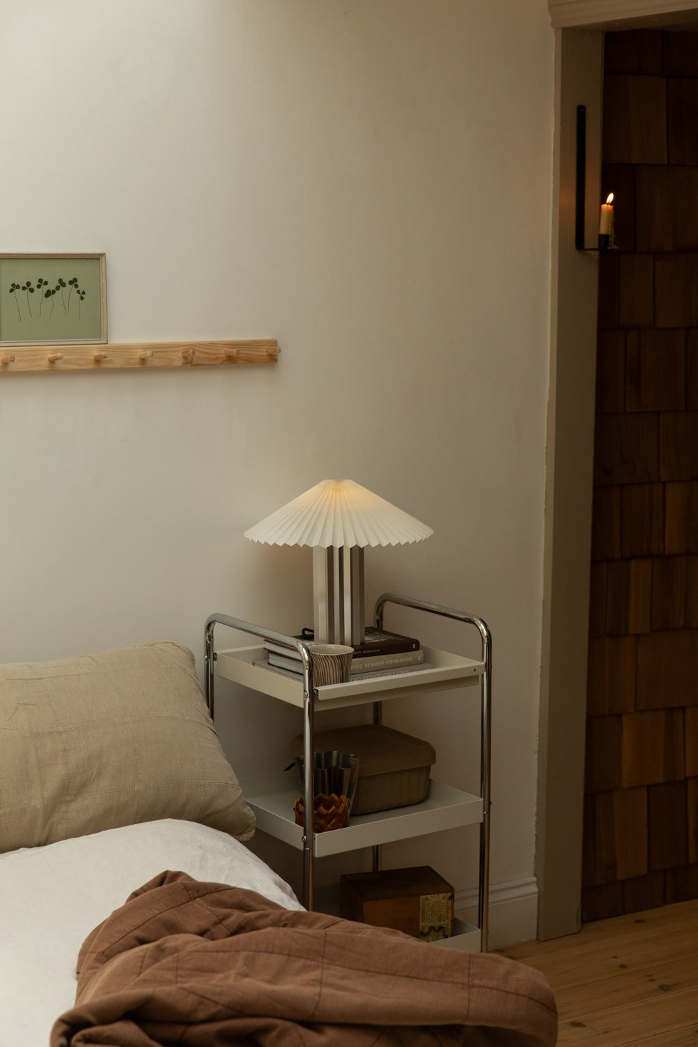 Blomus Taite lamp in lily white on chrome trolley nightstand beside linen bed, wall candle sconce glowing in timber-clad doorway.
