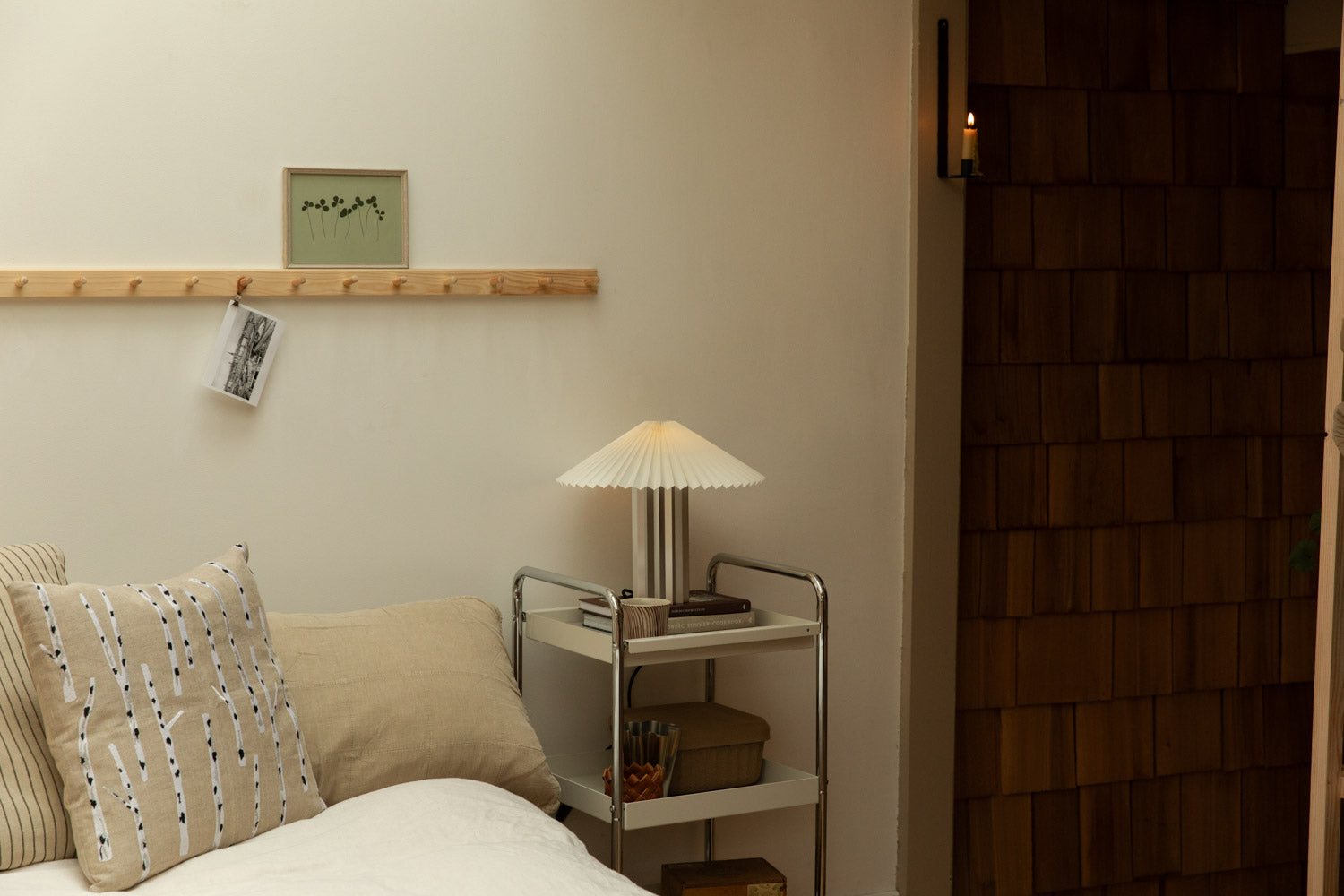 Blomus Taite lamp in lily white on chrome trolley nightstand beside linen bed with an embroidered birch cushion, wall candle sconce glowing in timber-clad doorway.