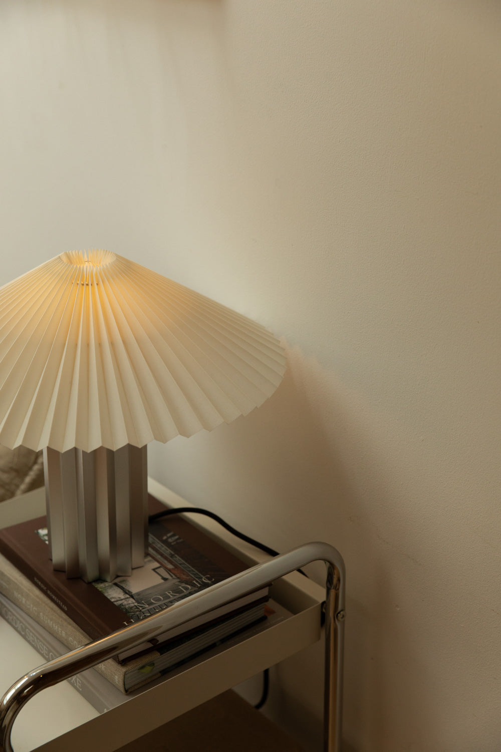Close-up of Blomus Taite lamp lily white pleated shade glowing warmly on chrome trolley with stacked Nordic books.