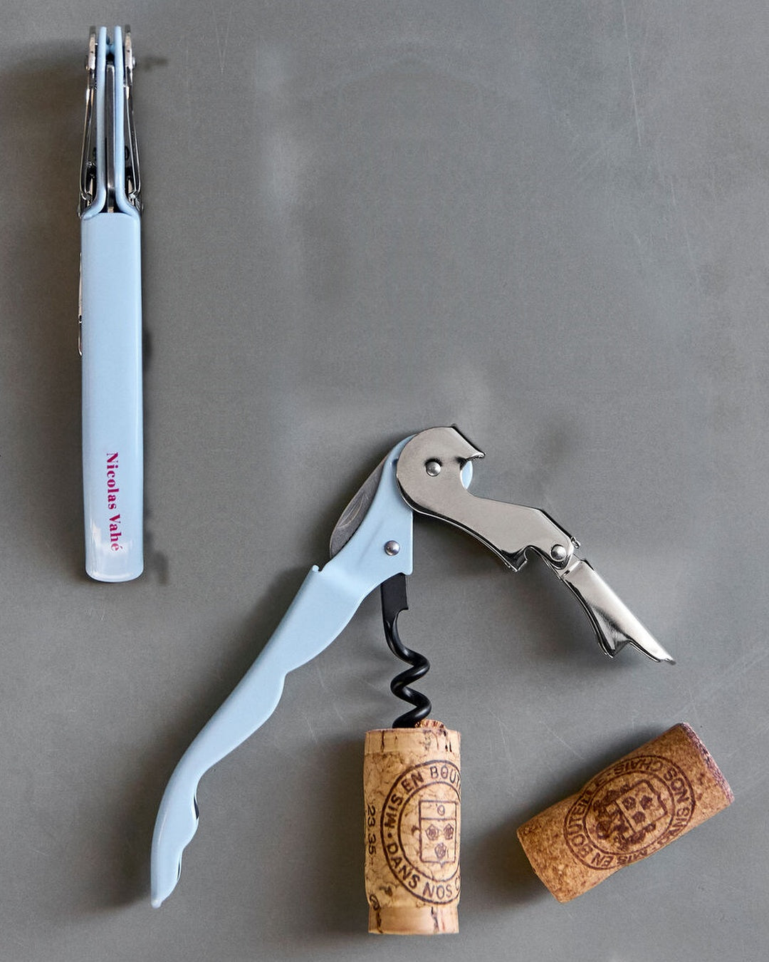 Light blue Nicolas Vahé bottle opener, open and closed, with corks on grey concrete