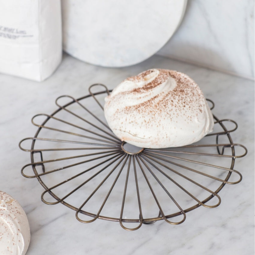 A brass cake rack with a circular base and curved wire supports, displaying a cake on top, placed on a marble surface.