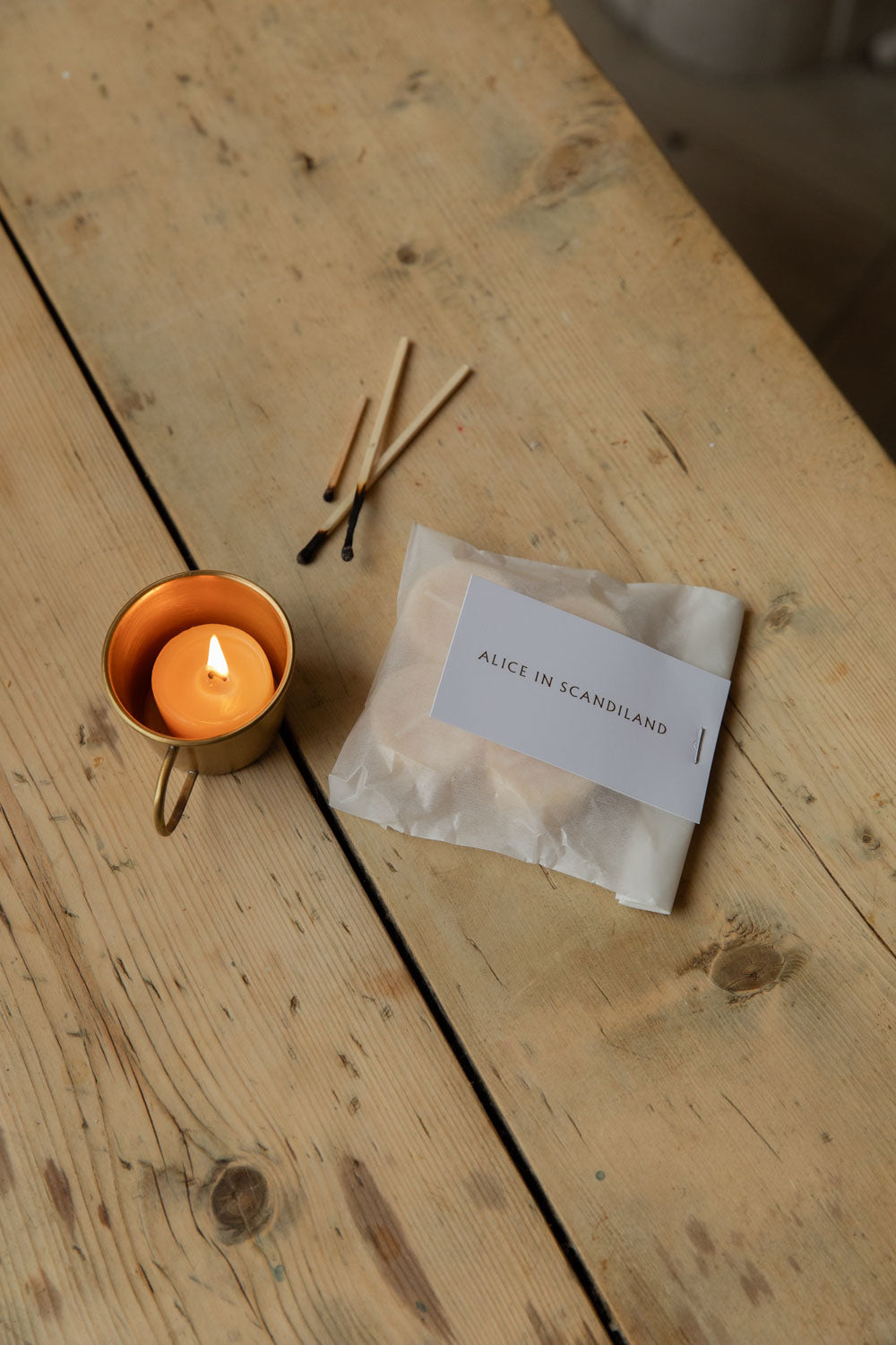 Lit beeswax tealight in a brass candle cup beside a labelled glassine pack and matches
