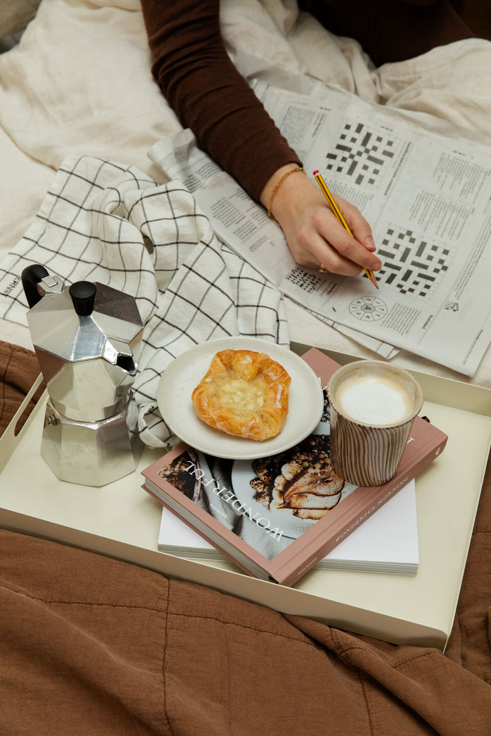 Woman doing a crossword in bed with a Bricka off-white tray with a Diva stripe mug, pastry, and Bialetti Moka Express.
