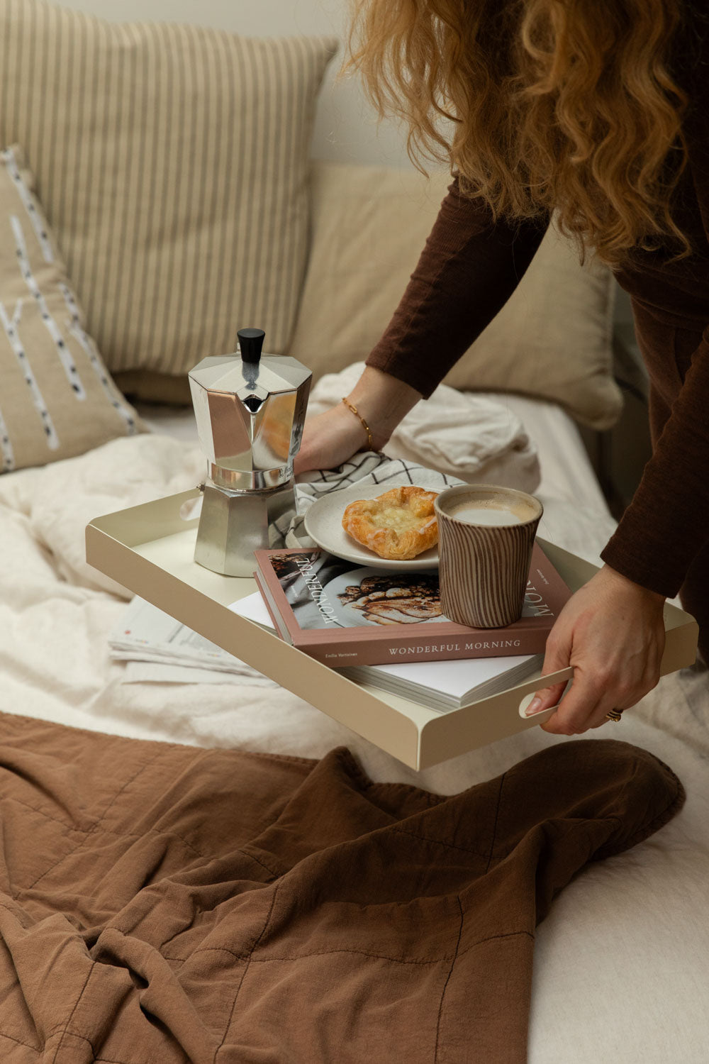 Woman holding a Bricka off-white tray with Diva stripe mug, pastry and Bialetti Moka Express on a bed with rust quilted throw and stripe cushions.