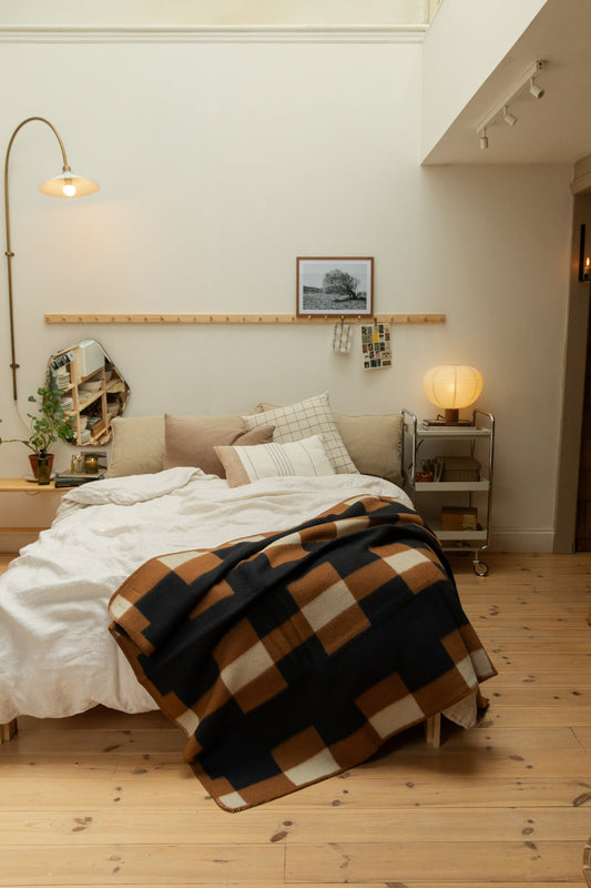 Brita Sweden Marta lambswool blanket in charcoal, tan and cream draped across a white linen bed, with brass floor lamp and pine peg rail.