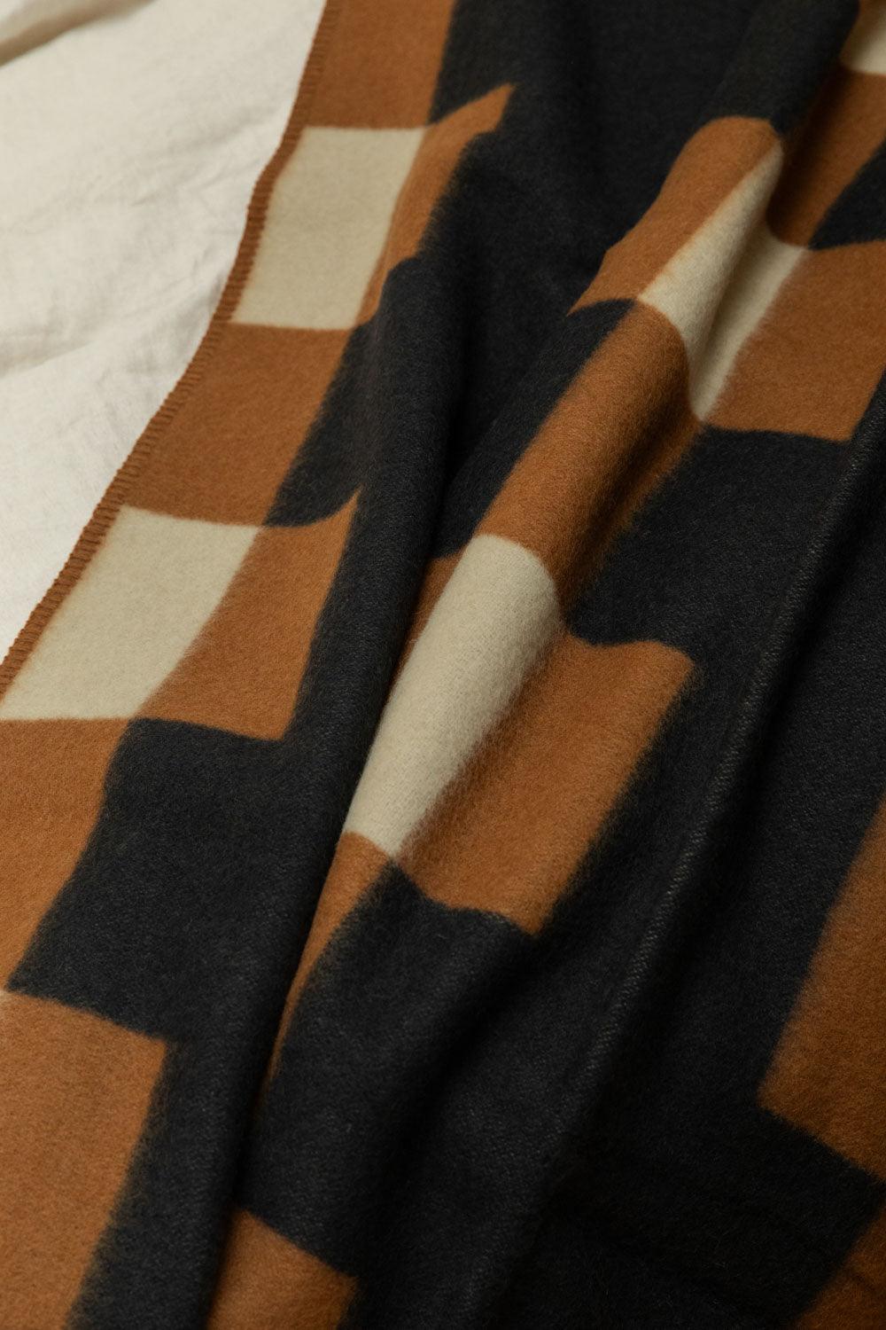 Close detail of Brita Sweden Marta lambswool blanket in charcoal, tan and cream geometric block pattern draped on a white linen bed.