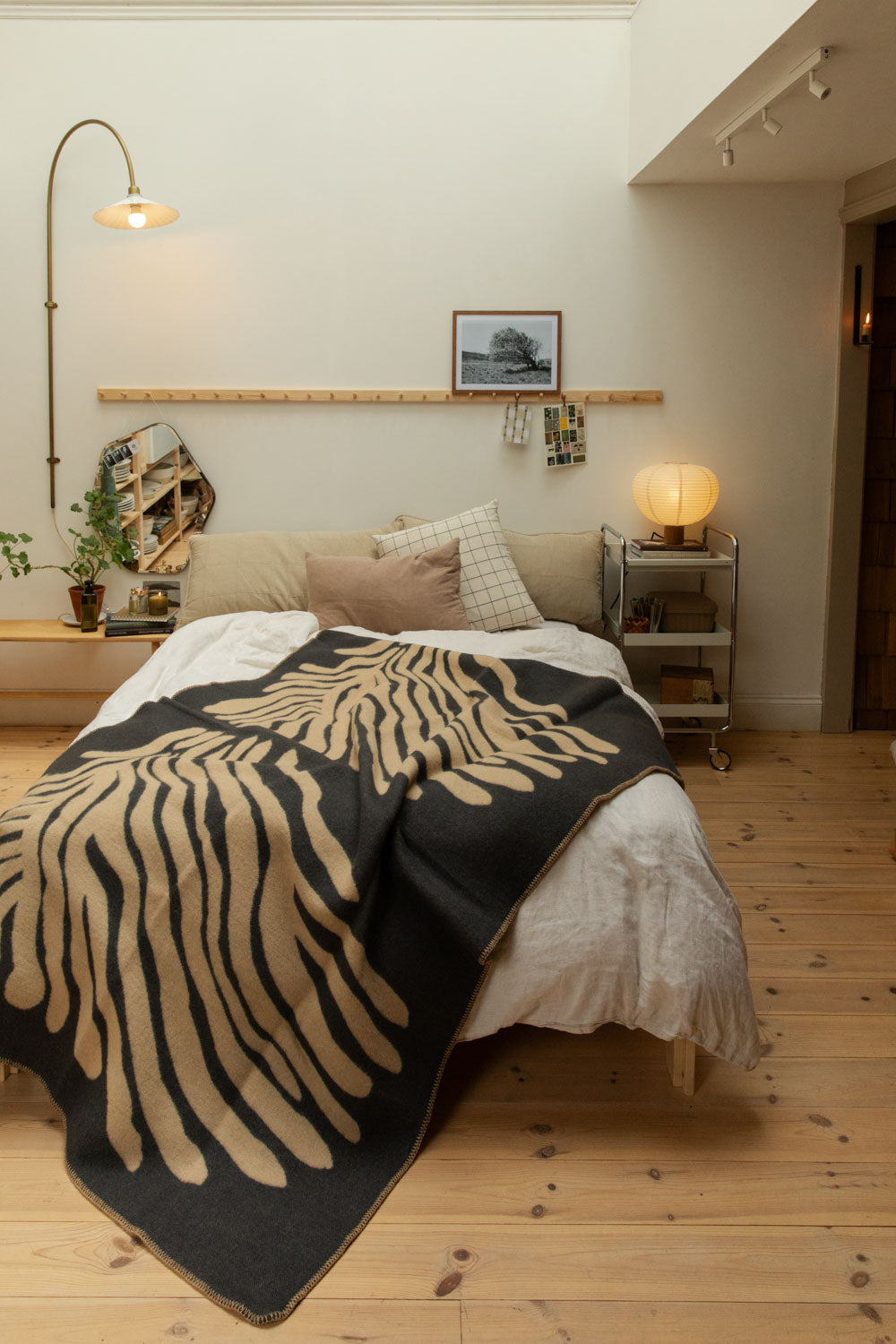 Full bedroom view with Brita Sweden Pine lambswool blanket in dark blue and sand on a bed with arc brass floor lamp, pine peg rail, paper lantern table lamp and pine floorboards.