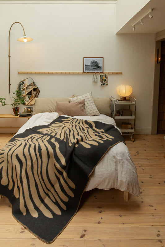 Full bedroom view with Brita Sweden Pine lambswool blanket in dark blue and sand on a bed with arc brass floor lamp, pine peg rail, paper lantern table lamp and pine floorboards.