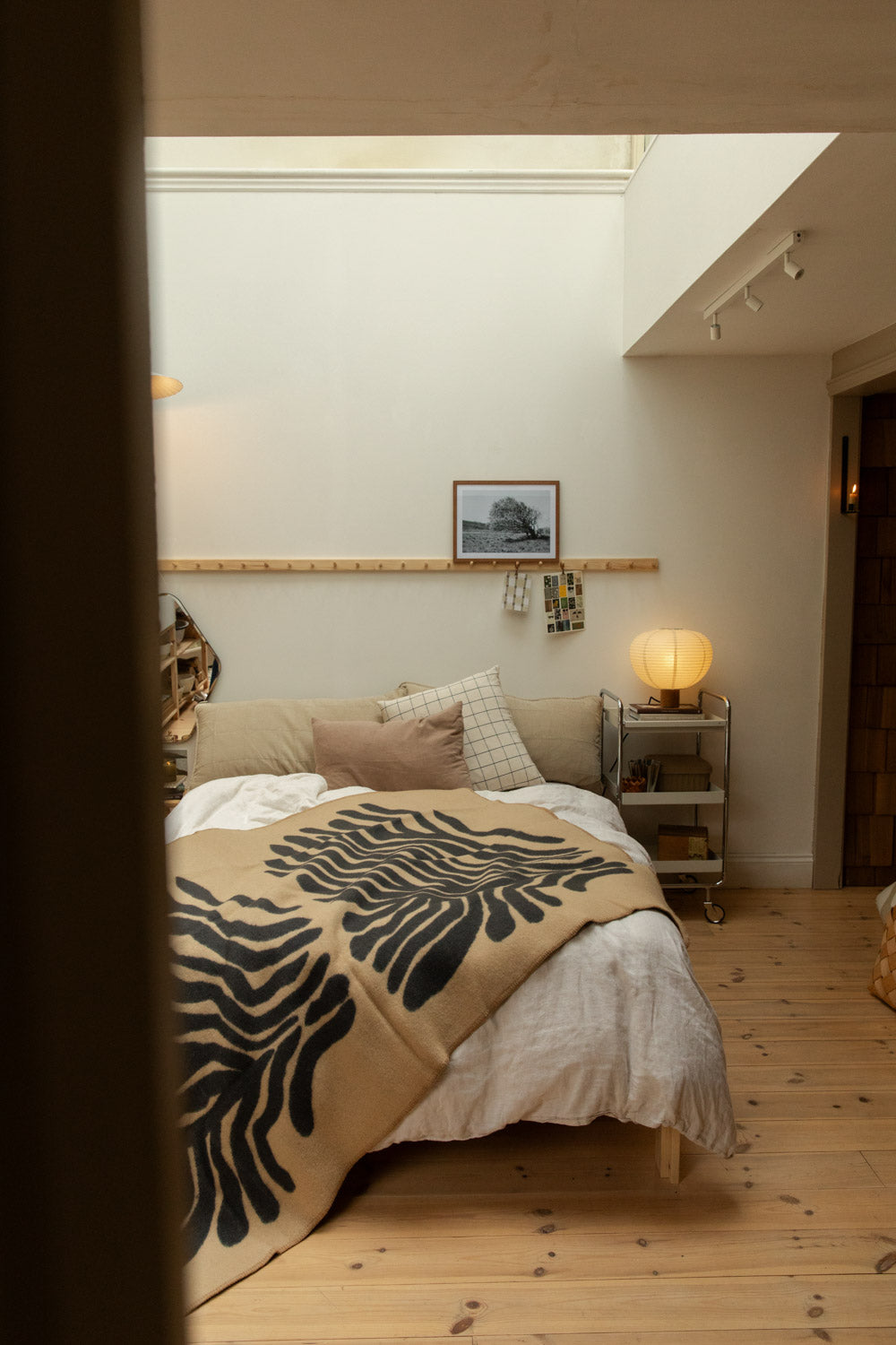 Full bedroom view with Brita Sweden Pine lambswool blanket in sand and dark blue draped over a bed with pine peg rail, paper lantern lamp and chrome trolley bedside table.