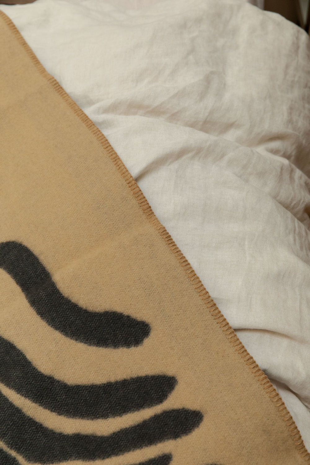 Close-up of Brita Sweden Pine lambswool blanket in sand and dark blue showing blanket stitch edge detail against white linen bedding.