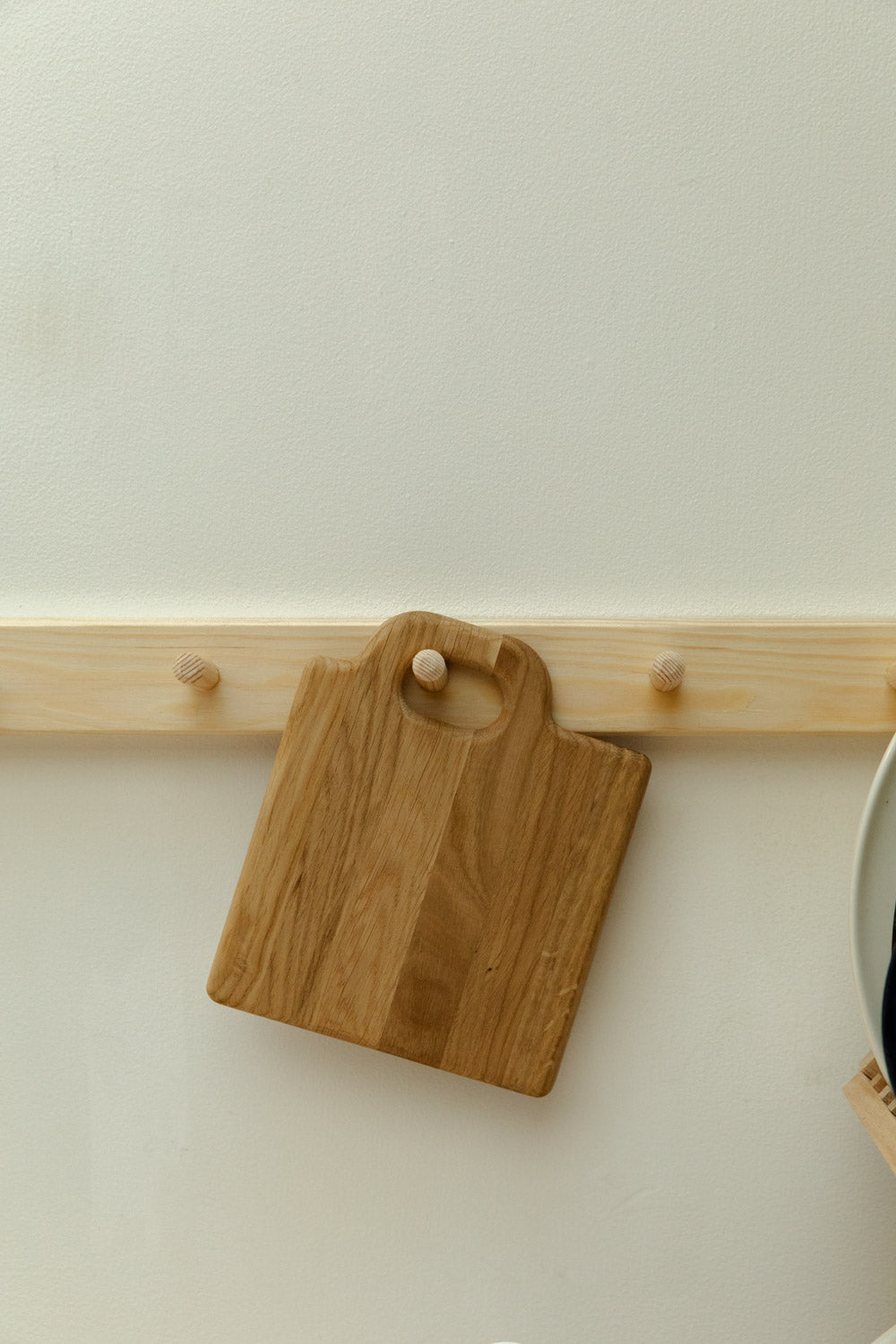 Small Broste Copenhagen Olina oak chopping board hung up on a wooden peg rail against a white background.