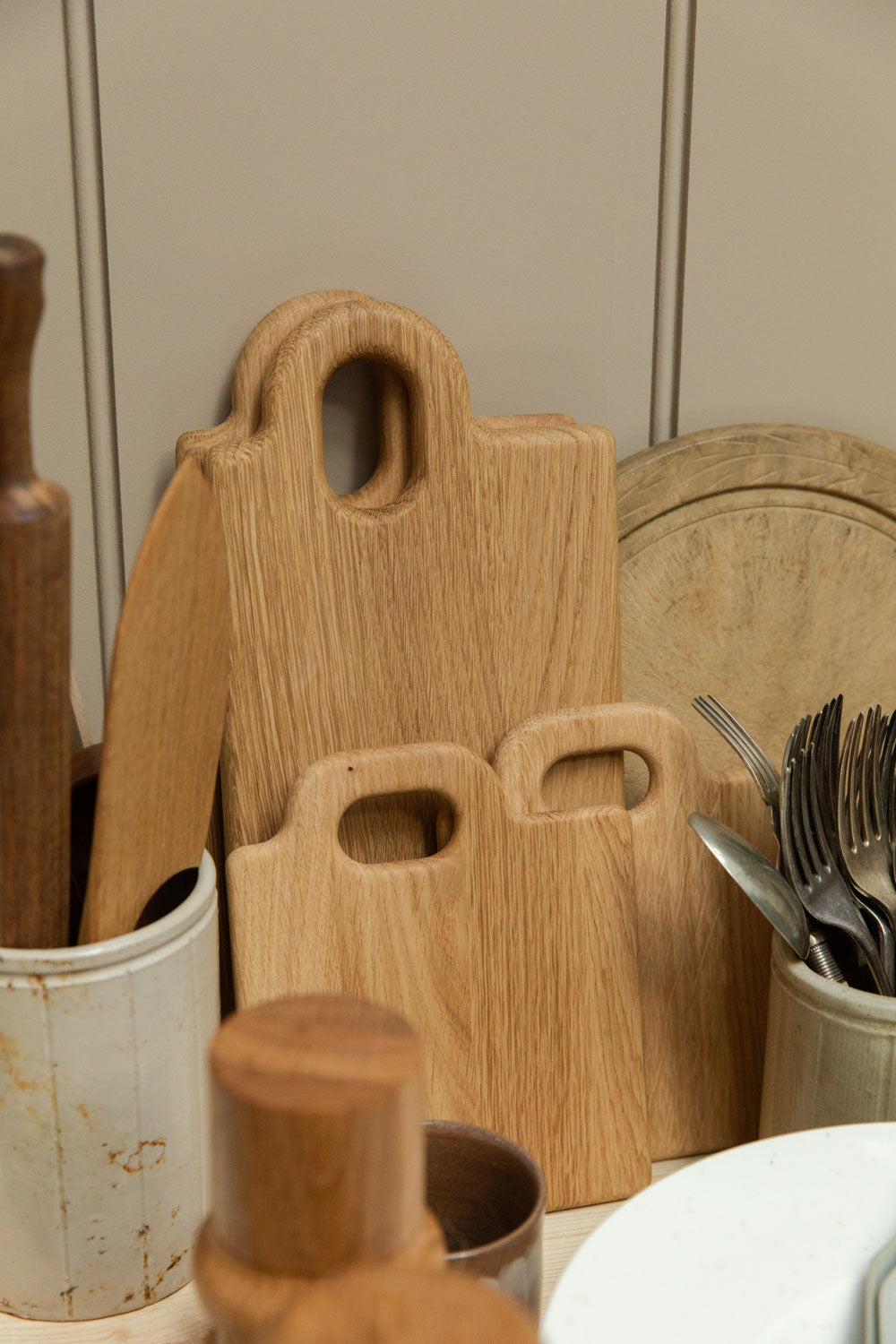Broste Copenhagen Olina oak chopping boards in two sizes leaning against taupe panelled wall, styled with vintage cutlery pot, rolling pin and round wooden board.