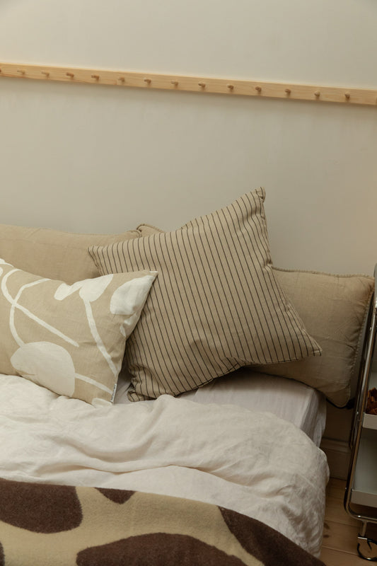 Broste Kare stripe cushion cover styled on a linen headboard with a floral print cushion, white linen duvet and Brita Sweden blanket at foot of bed.