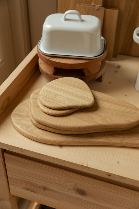 Stacked Broste Limfjord oak chopping boards in three sizes on a pine kitchen table, with white enamel butter dish on wooden riser behind