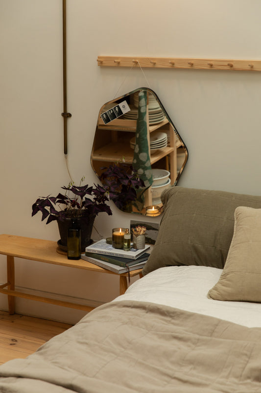 Broste Ruby Liten organic shaped mirror hanging above a pine bench bedside table styled with Nordic books, lit candle, perfume bottle and purple oxalis plant, with eucalyptus and fog quilted linen headboard cushions on a white linen bed.