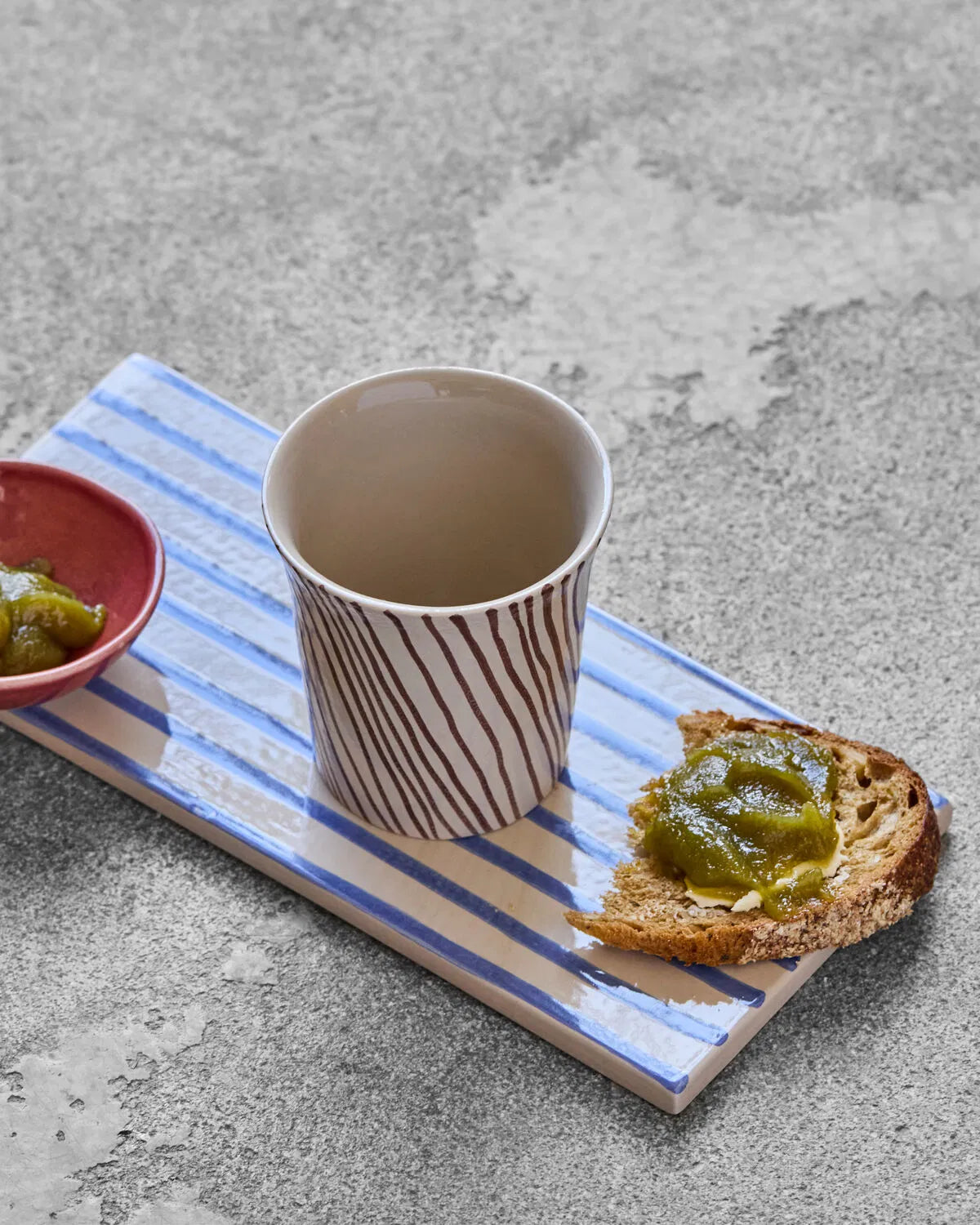 A brown striped Diva mug is placed on a blue and white striped tile setting, on top of a concrete background, with a piece of toast with green relish placed next to it.