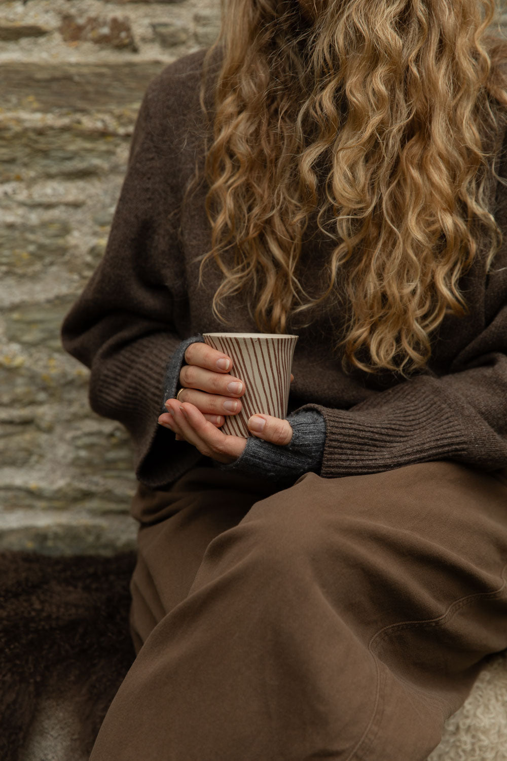 Cream stoneware cup with fine brown diagonal stripes held in hands over a brown skirt