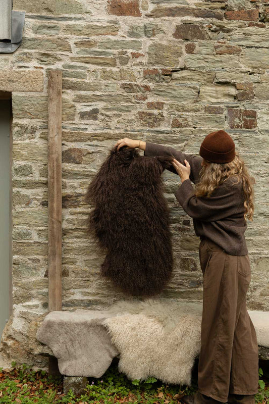 Long-haired chocolate-brown Tibetan sheepskin rug held against a stone wall, a cream sheepskin on the bench below