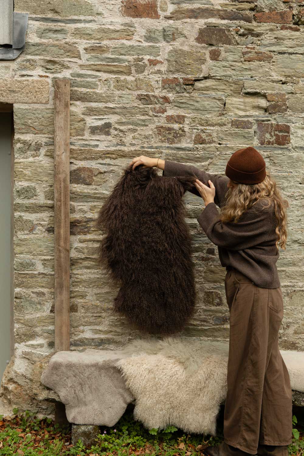 Long-haired chocolate-brown Tibetan sheepskin rug held against a stone wall, a cream sheepskin on the bench below