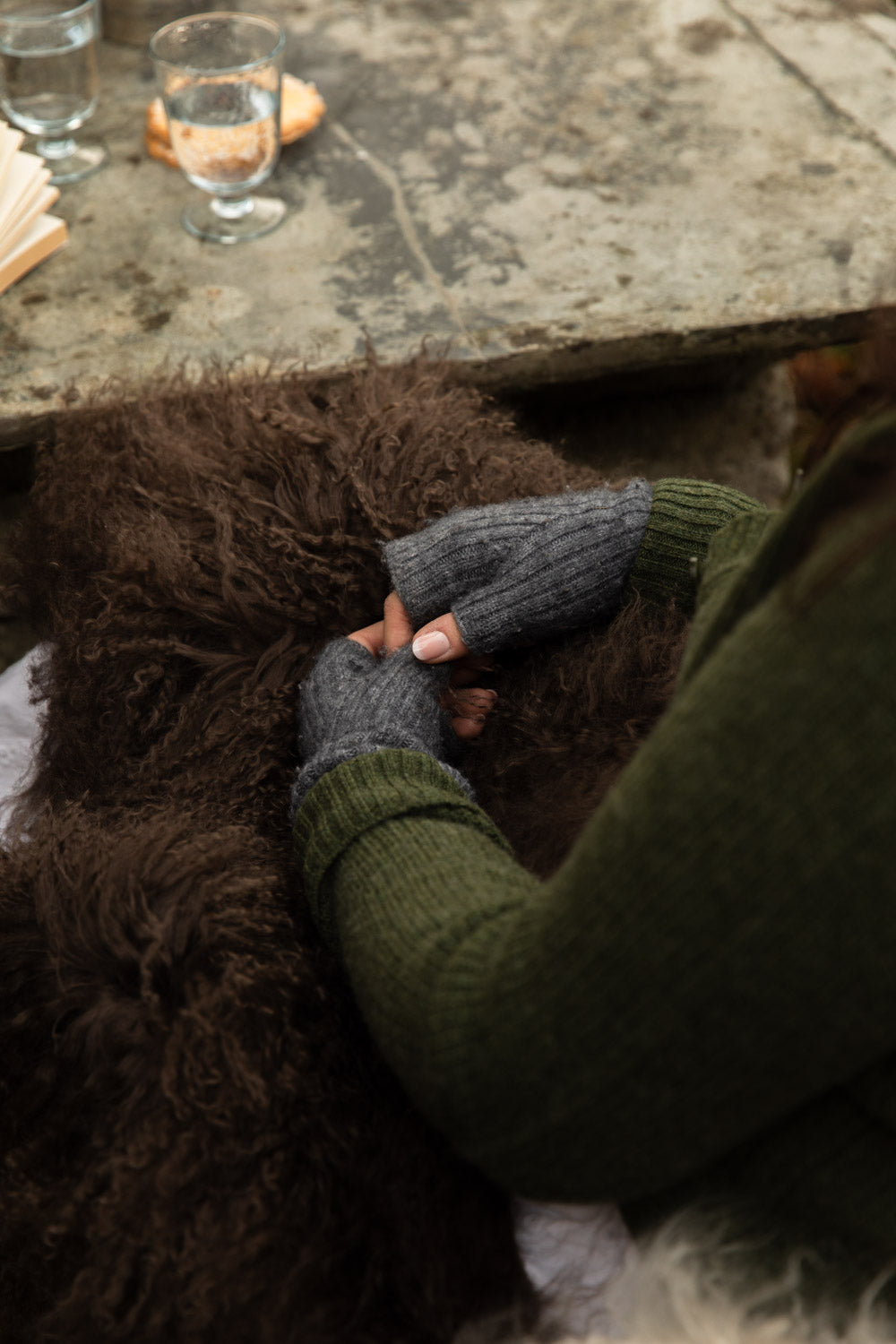 Hands in grey knitted wrist warmers resting on a long, curly chocolate-brown Tibetan sheepskin rug at an outdoor stone table