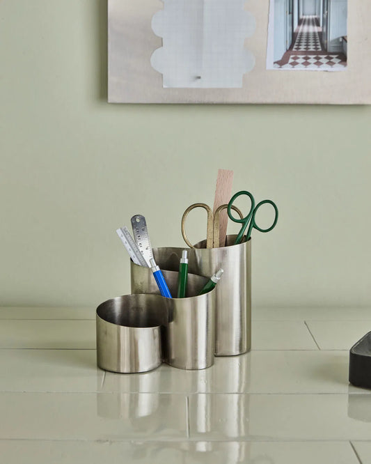Brushed silver curve organiser placed on a light green tiled surface, against a background of a similar colour. The organiser is filled with scissors, rules and pens. 